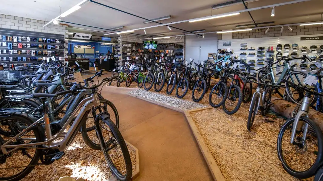 Innenraum eines Fahrradladens mit vielen Mountainbikes und E-Bikes auf Holzspänen neben einem Werkstattbereich.