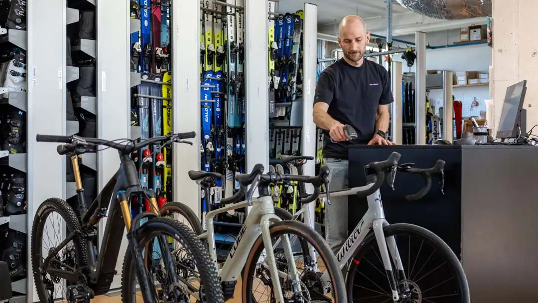 Mann scannt an der Kasse in einem Sportgeschäft mit Fahrrädern und Skiausrüstung im Hintergrund.