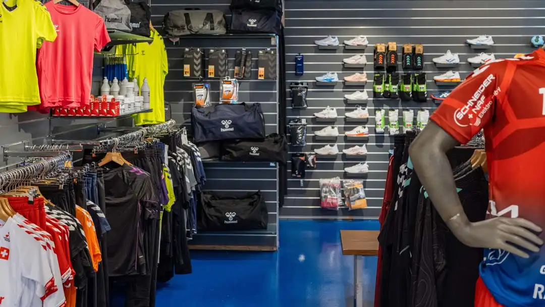 Sportgeschäft mit Sportbekleidung, Taschen, Schuhen und Accessoires an Regalen und einem Schaufenstermännchen.