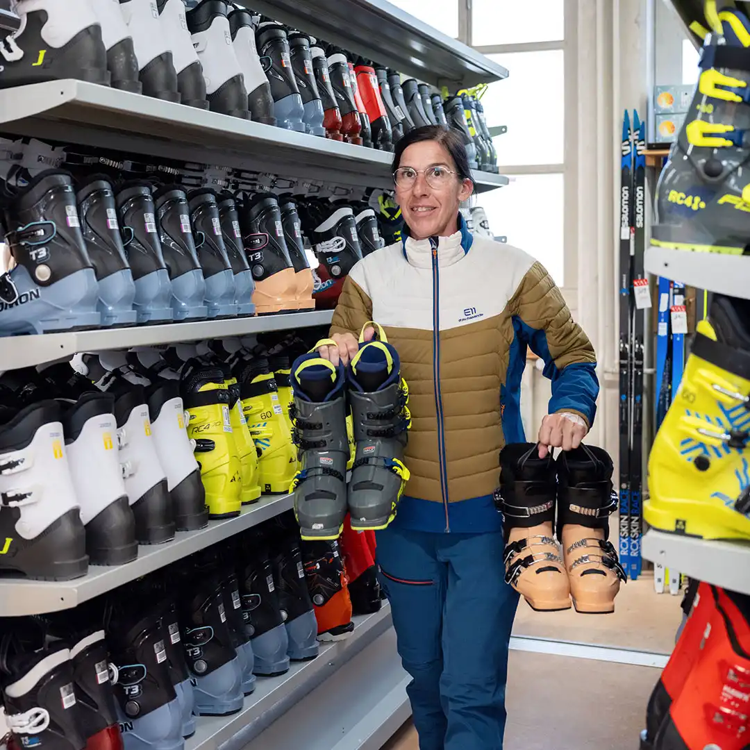 Frau in Jacke zeigt Skischuhe im Verleih mit Regalen voller Skischuhe.