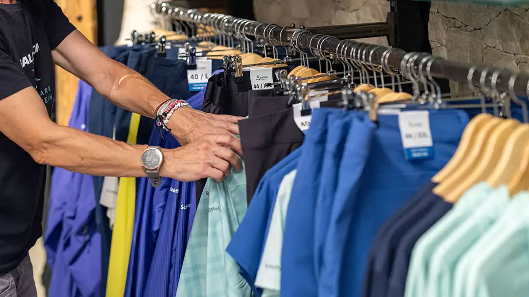 Person tastet in einem Geschäft Kleidung auf einer Stange mit blauen und schwarzen Hosen ab.