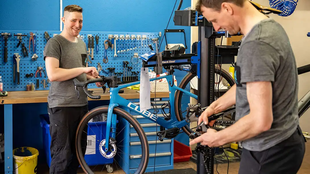 Zwei Männer reparieren Fahrräder in einer Werkstatt mit Werkzeugwand im Hintergrund.