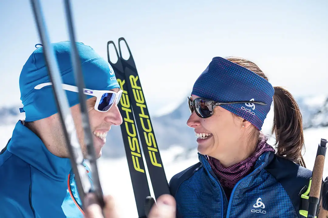 Zwei Skilangläufer in blauer Sportkleidung lachen und halten Skier der Marke Fischer.