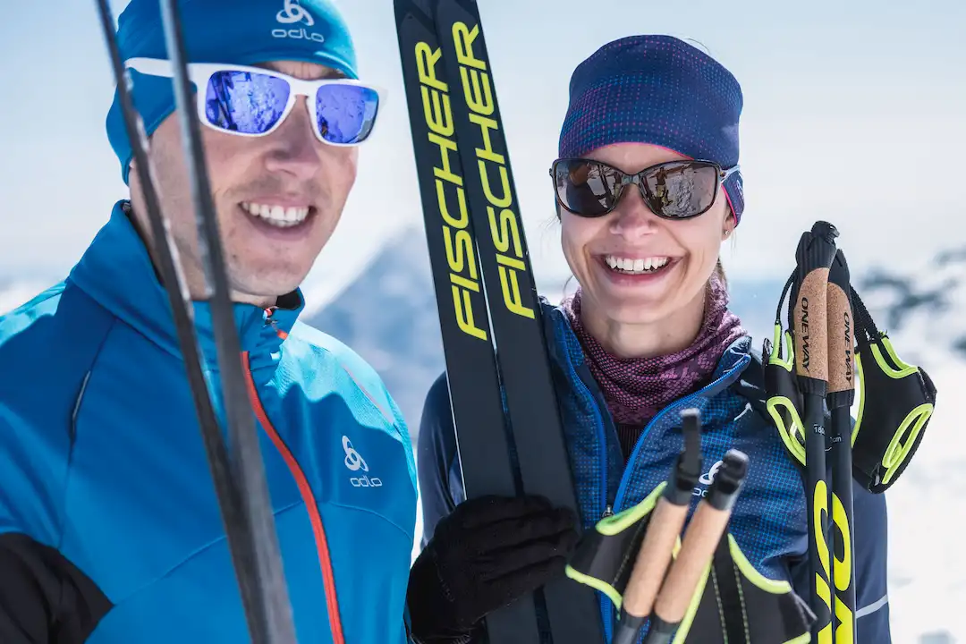 Zwei lächelnde Langläufer in blauer Winterkleidung mit Skiern und Stöcken vor Bergkulisse.