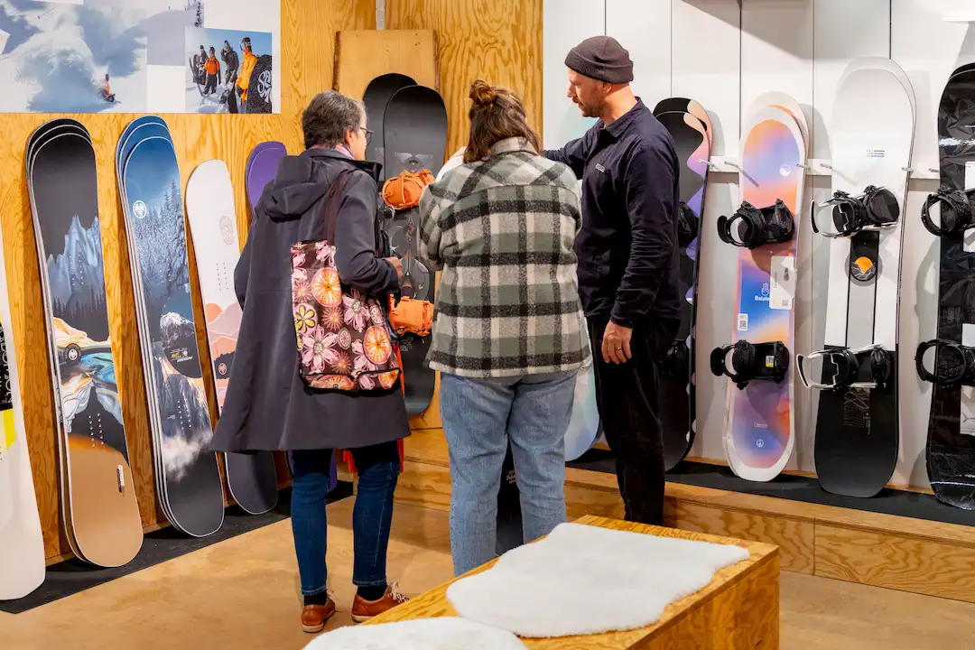 Drei Personen betrachten und besprechen Snowboards in einem Geschäft mit Wanddisplay.