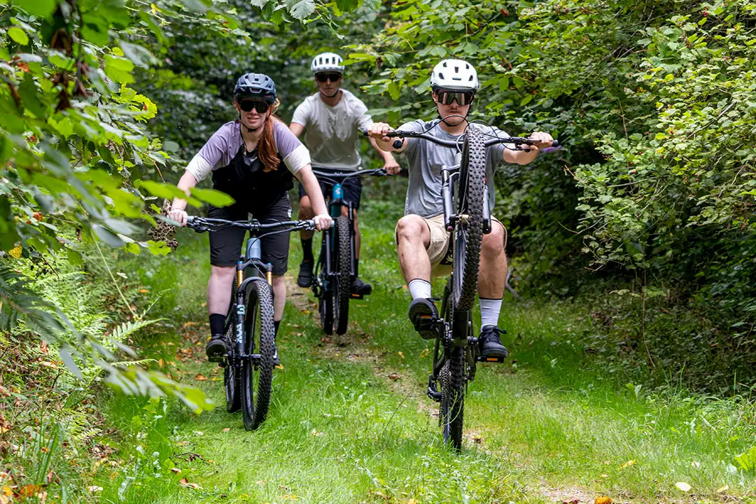 Drei Personen fahren Mountainbike auf einem bewachsenen Waldweg, vorn einer macht ein Wheelie.