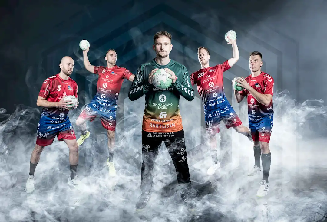 Fünf Handballspieler in Action, einer in grünem Trikot im Zentrum, umgeben von Nebel.