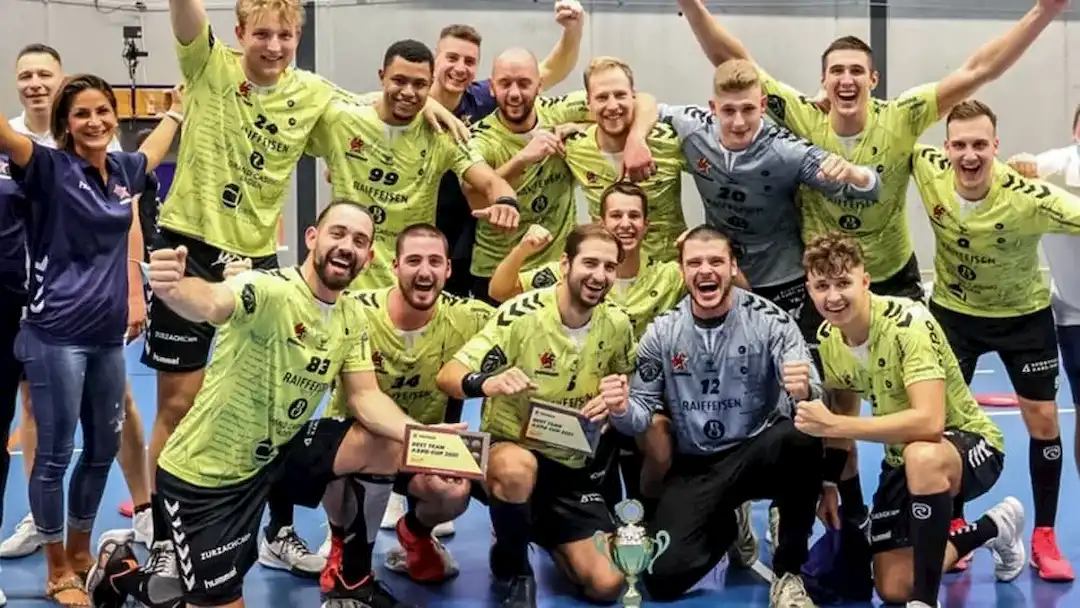 Jubelndes Handballteam mit Pokal und Auszeichnungen auf der Sporthalle, Spieler tragen gelbe Trikots.