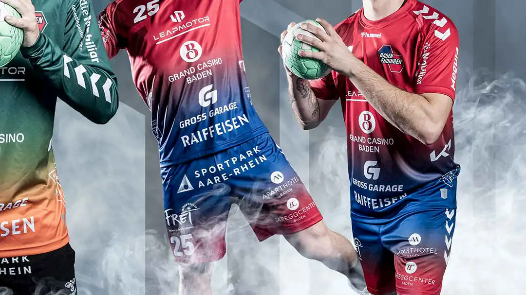 Zwei Handballspieler in rot-blauem Trikot mit Ball, Nebel im Hintergrund.