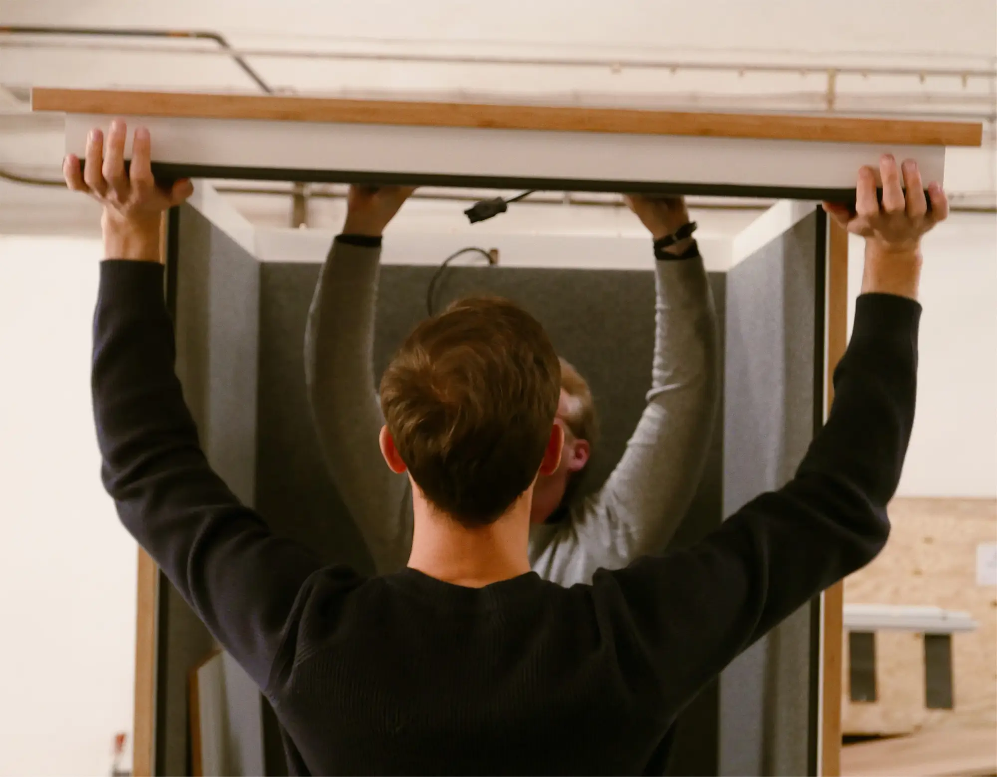 Two men assembling a booth or small enclosure, lifting a panel to place on top.