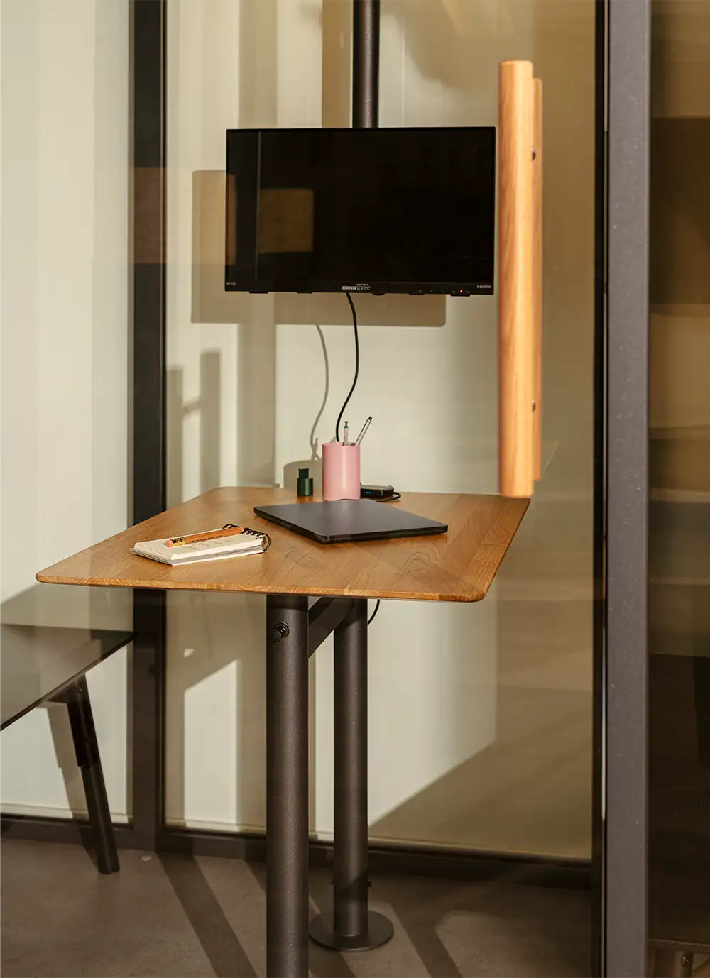 Minimalist office workspace with a wooden desk, closed laptop, notebook, pen holder, and wall-mounted monitor.