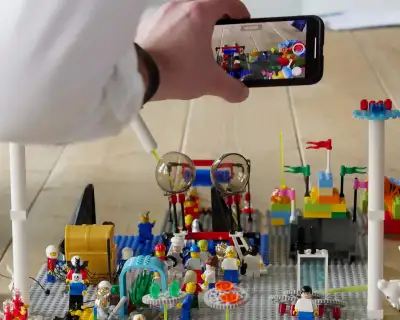 Foto einer Person, die ein Smartphone hält und ein Video von einer Lego-Anlage aufnimmt, die verschiedene Figuren und Strukturen auf einer flachen Oberfläche zeigt. Auf dem Smartphone-Bildschirm ist eine Nahaufnahme der Lego-Szene zu sehen, mit bunten Blöcken und Miniaturfiguren in Aktion. Zwei große transparente Kugeln und mehrere Lego-Figuren sind in der Anlage sichtbar.