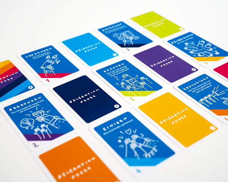 Ein Deck Delegation Poker Karten von HelloAgile in verschiedenen Farben liegt auf einem weißen Hintergrund ausgebreitet. Die Karten enthalten Illustrationen und Begriffe auf Deutsch, die verschiedene Aspekte der Delegation und Entscheidungsfindung darstellen, wie "Begründen," "Einigen," und "Erkunden."