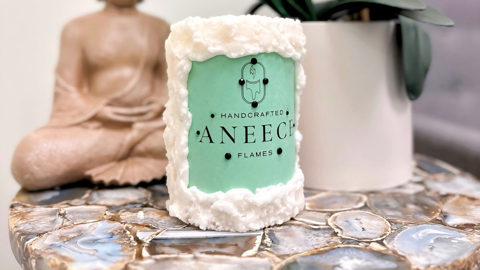 A handcrafted Aneece candle with the brand's logo set into the wax, beside a small Buddha figure and a potted orchid