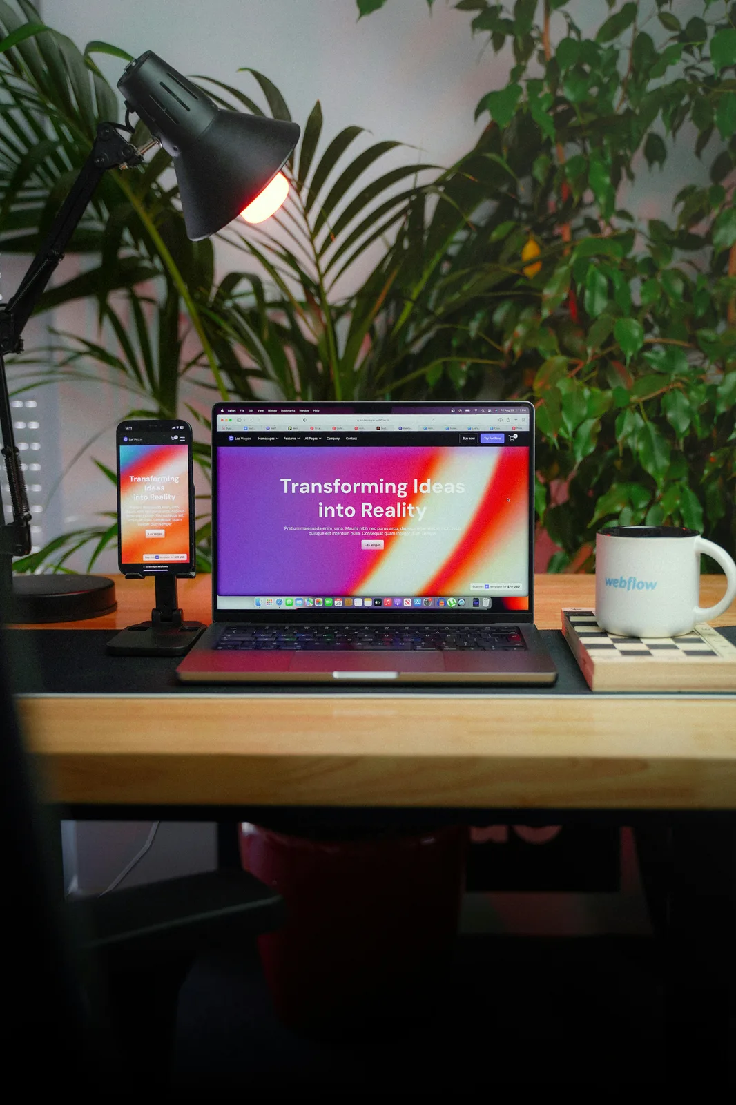 a laptop with a smartphone and mug with the Webflow logo on a wood desk in an office filled with plants in the background