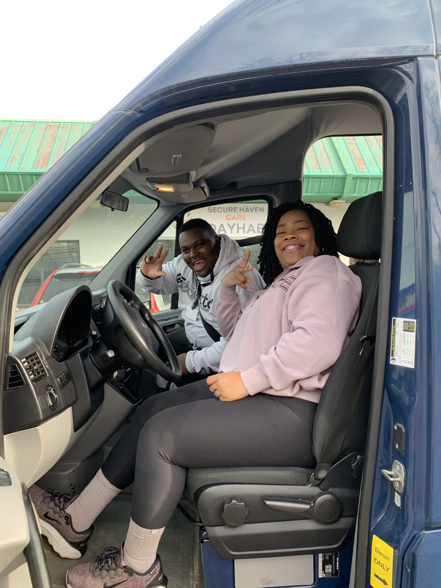 Two smiling people sitting inside a blue van, with the driver and front passenger seats visible.