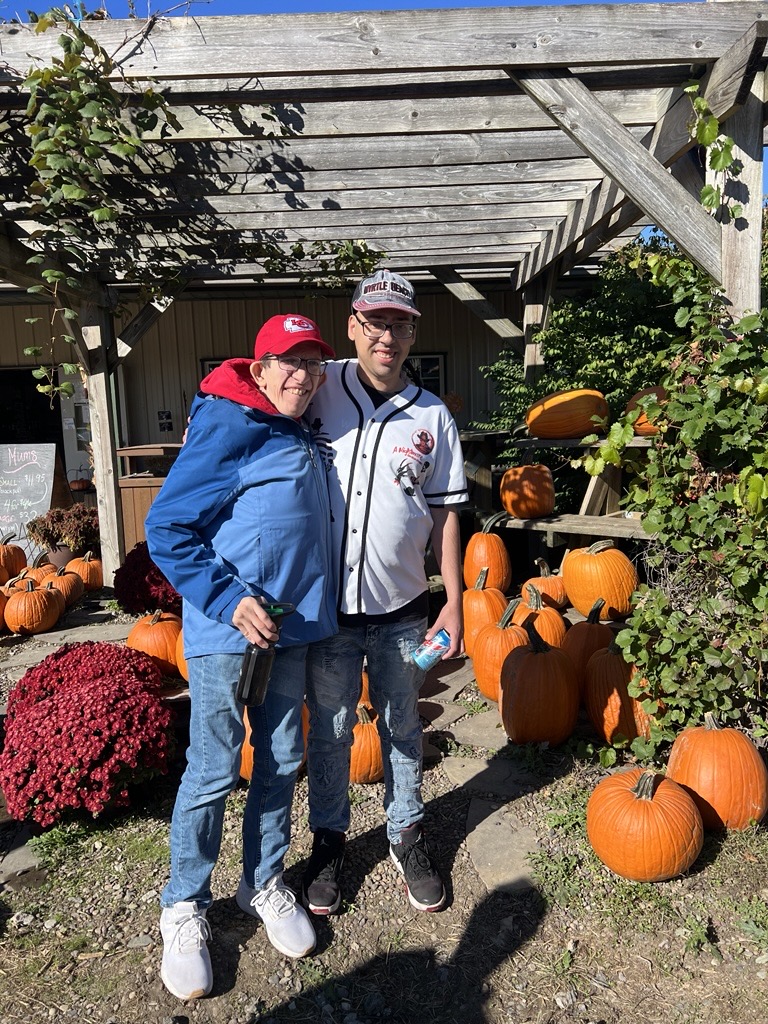 Two people smiling and hugging under a wooden pergola surrounded by pumpkins and autumn plants.
