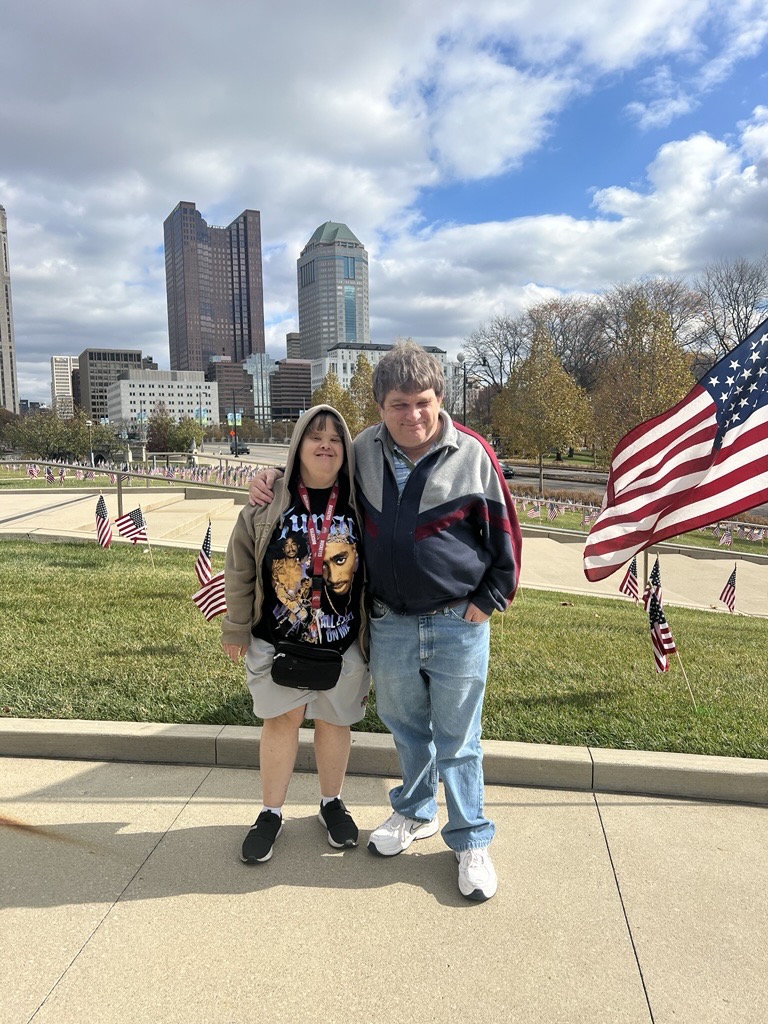 Two people standing arm in arm on a sidewalk with a city skyline and American flags in the background.