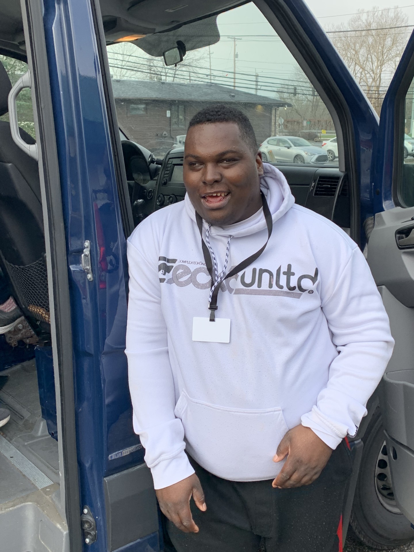 Smiling man wearing a white hoodie with a badge on a lanyard, standing in the doorway of a blue vehicle.