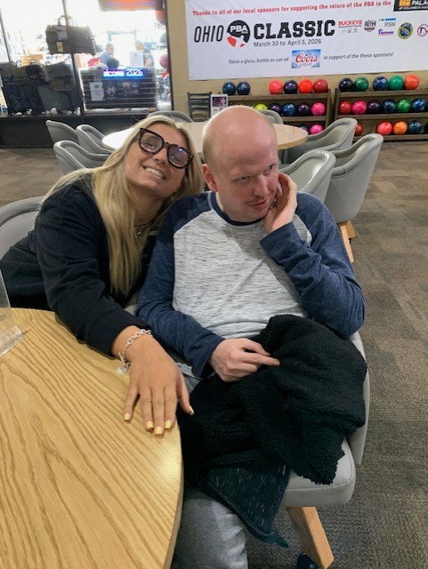 A woman with glasses smiling next to a man seated in a chair, in a bowling alley with colorful bowling balls in the background.