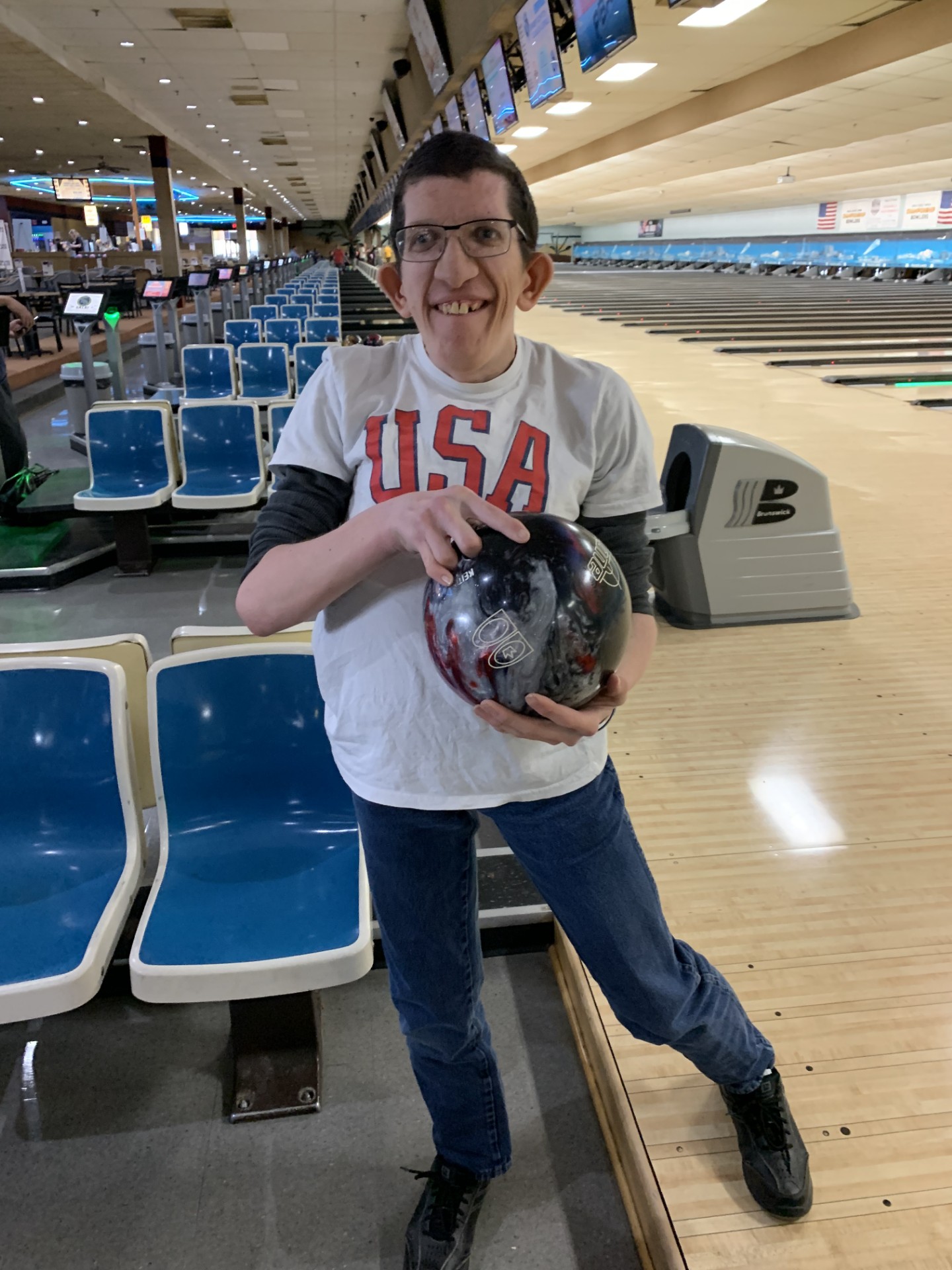 Person wearing glasses and a white USA t-shirt holding a bowling ball at a bowling alley.