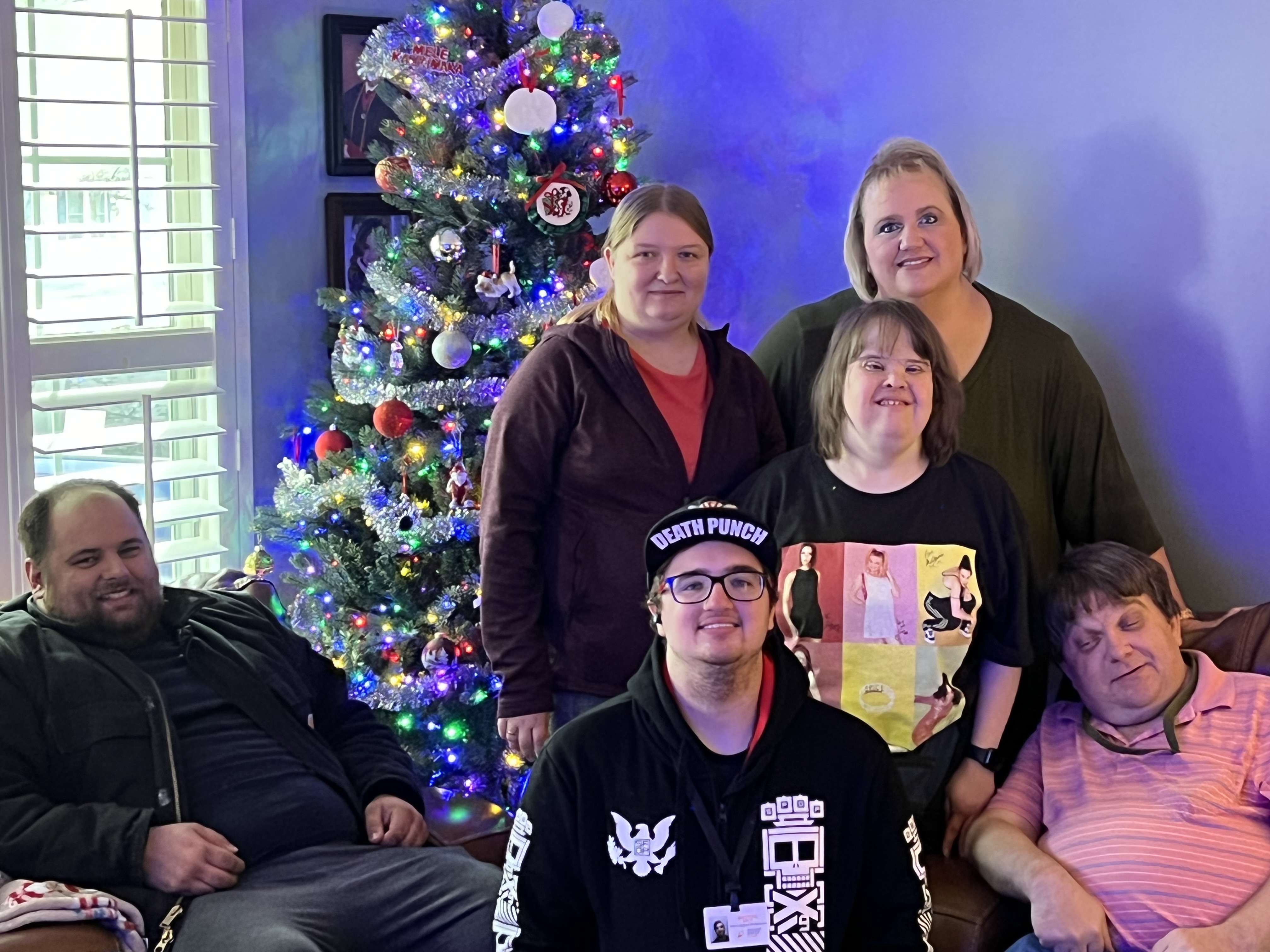 Group of six people smiling and posing in front of a decorated Christmas tree indoors.