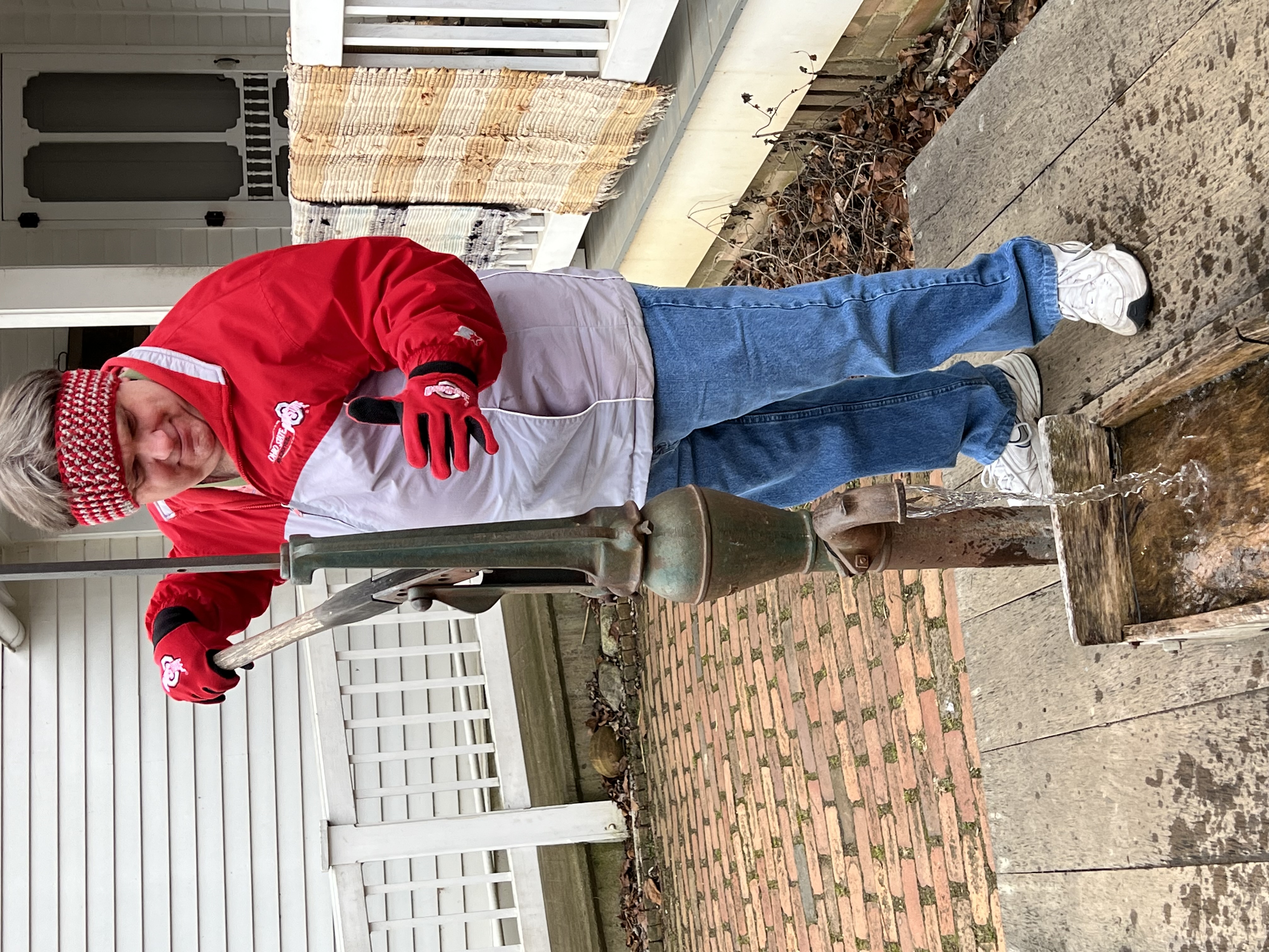 Person wearing red Ohio State jacket and gloves operating an antique water pump with water flowing into a wooden trough.