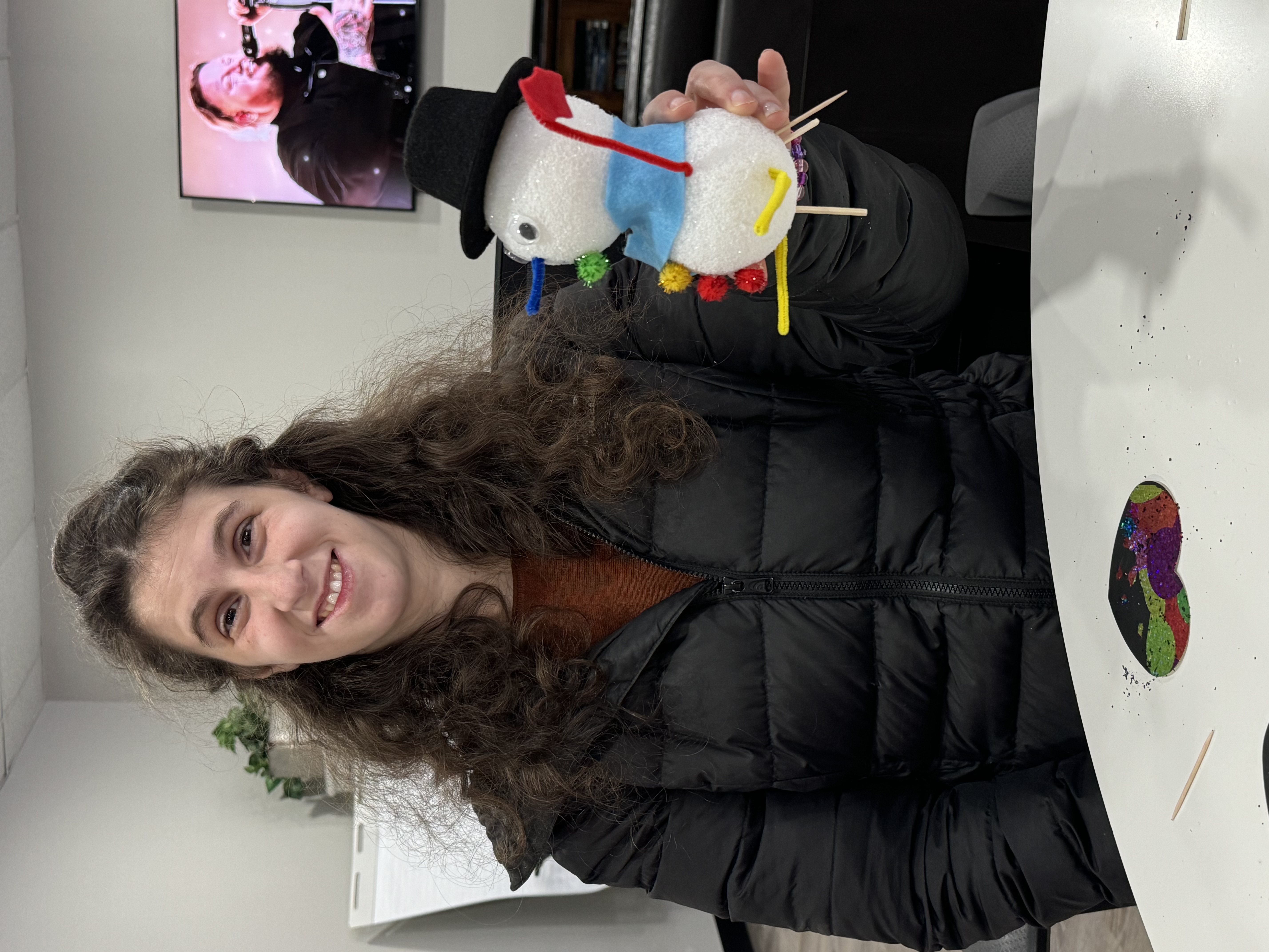 Smiling woman with curly hair wearing a black jacket, holding a craft snowman made of white foam balls decorated with a black hat, blue scarf, pom-poms, and pipe cleaner arms.