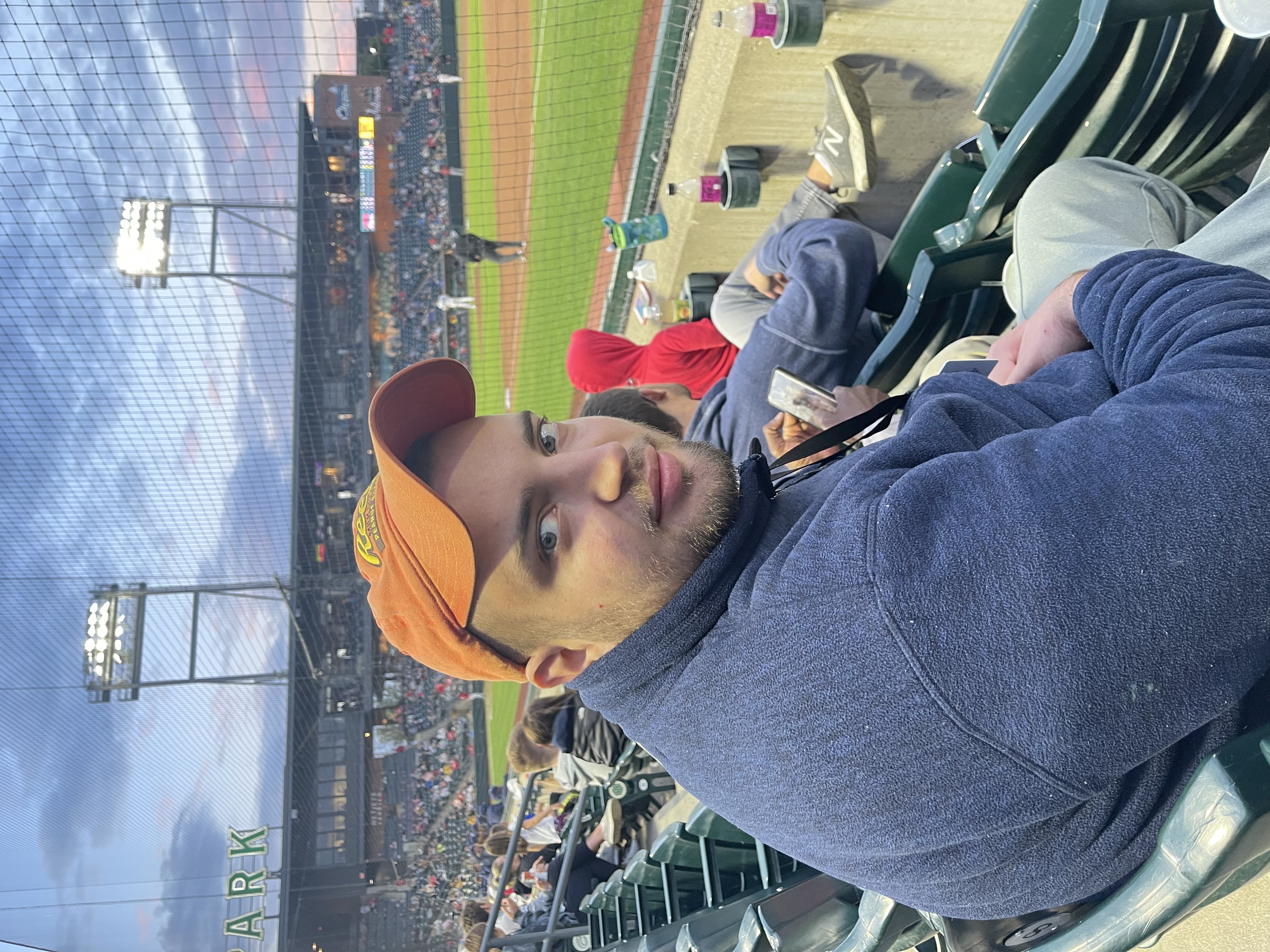 Man in an orange cap and blue jacket sitting in the stands at a baseball game during dusk, with the field and other spectators visible.