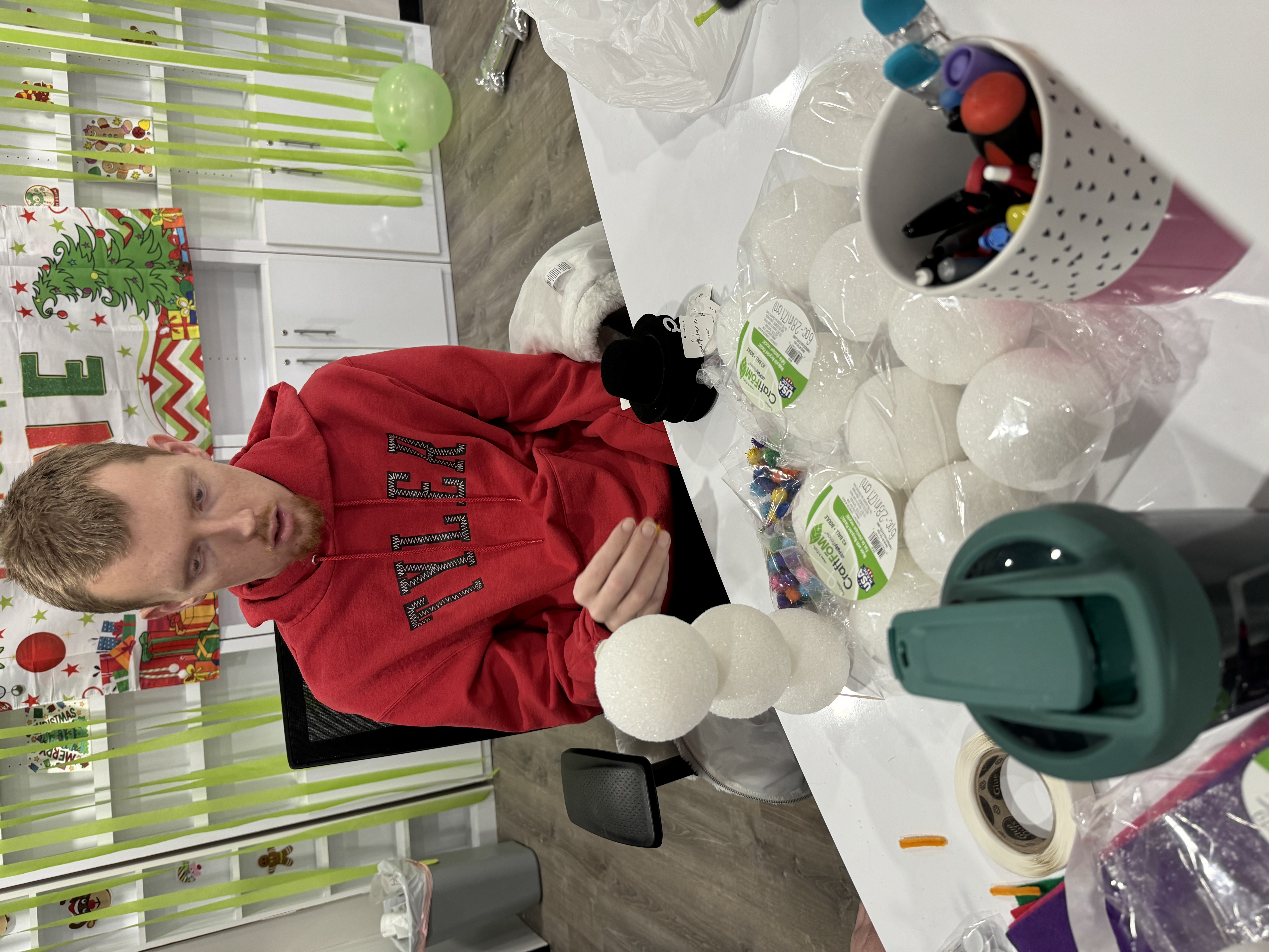 A person in a red hoodie assembling a snowman craft using three stacked white foam balls on a table with crafting supplies and decorations in the background.