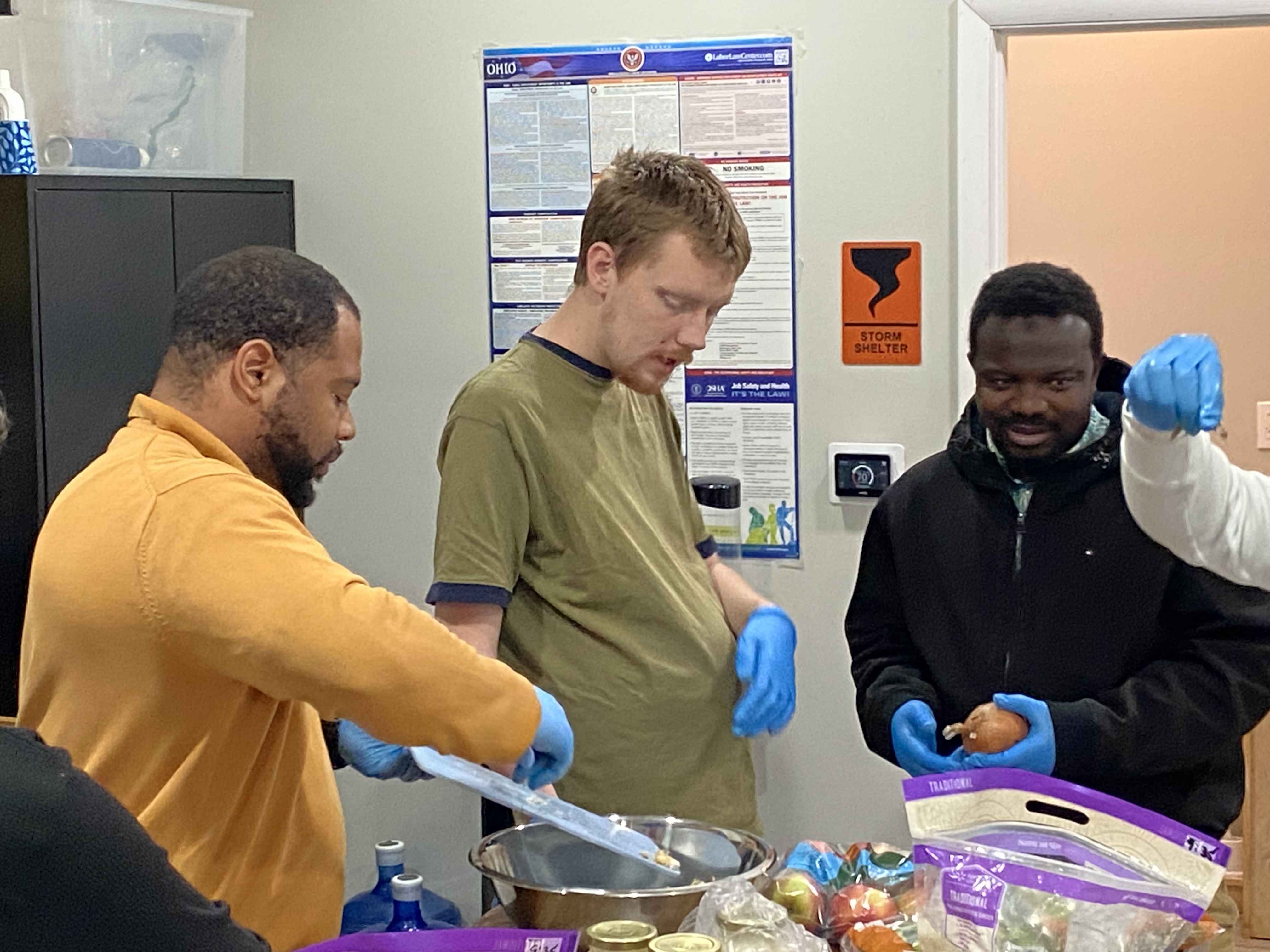 Three men wearing blue gloves prepare food in a kitchen, handling utensils and an onion near a bowl and bags of produce.