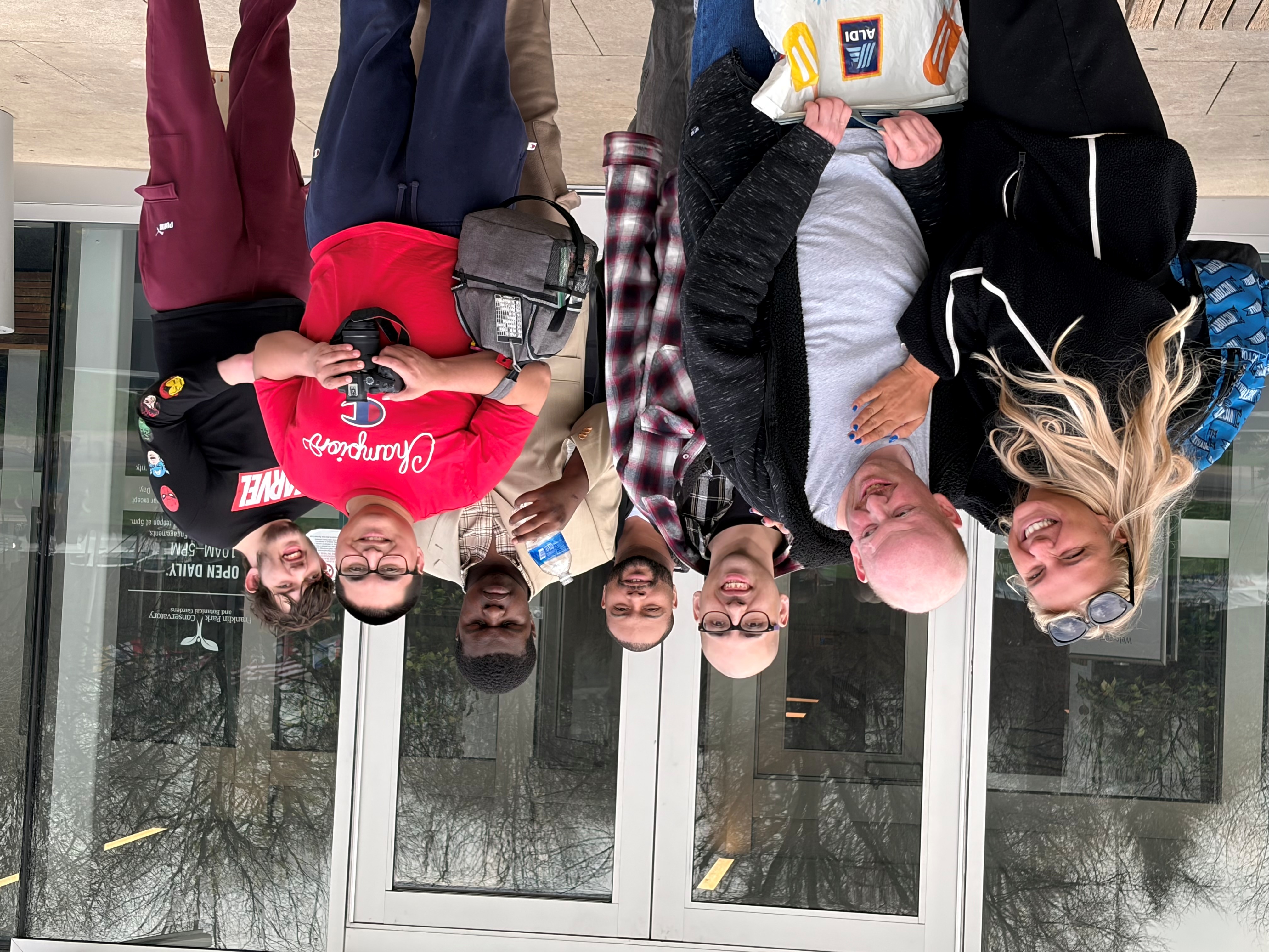 Group of seven diverse people standing closely and smiling in front of a glass door entrance to Franklin Park Conservatory.