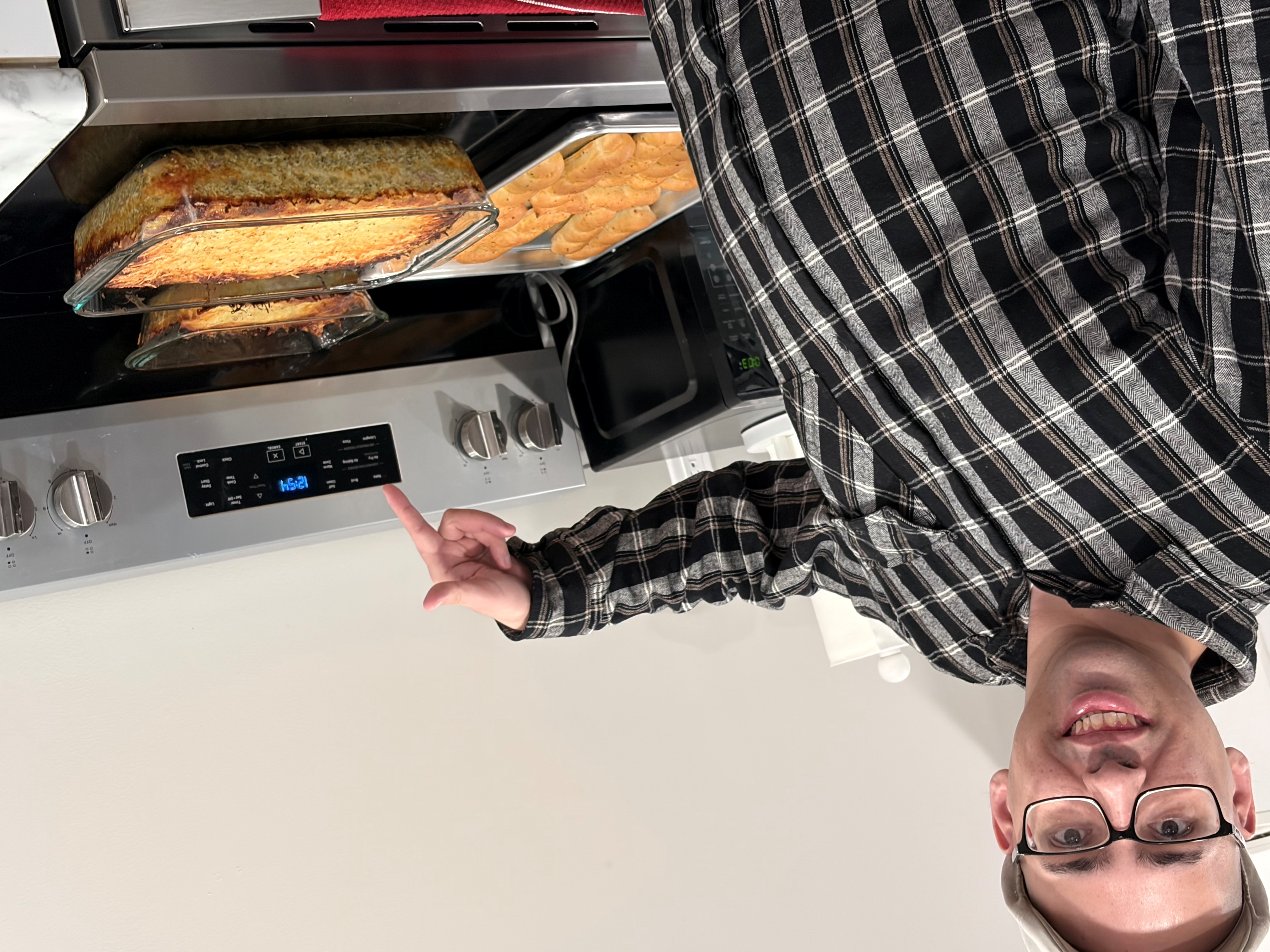 Smiling man wearing glasses and a cap points at an oven with baked dishes and rolls on the stove.
