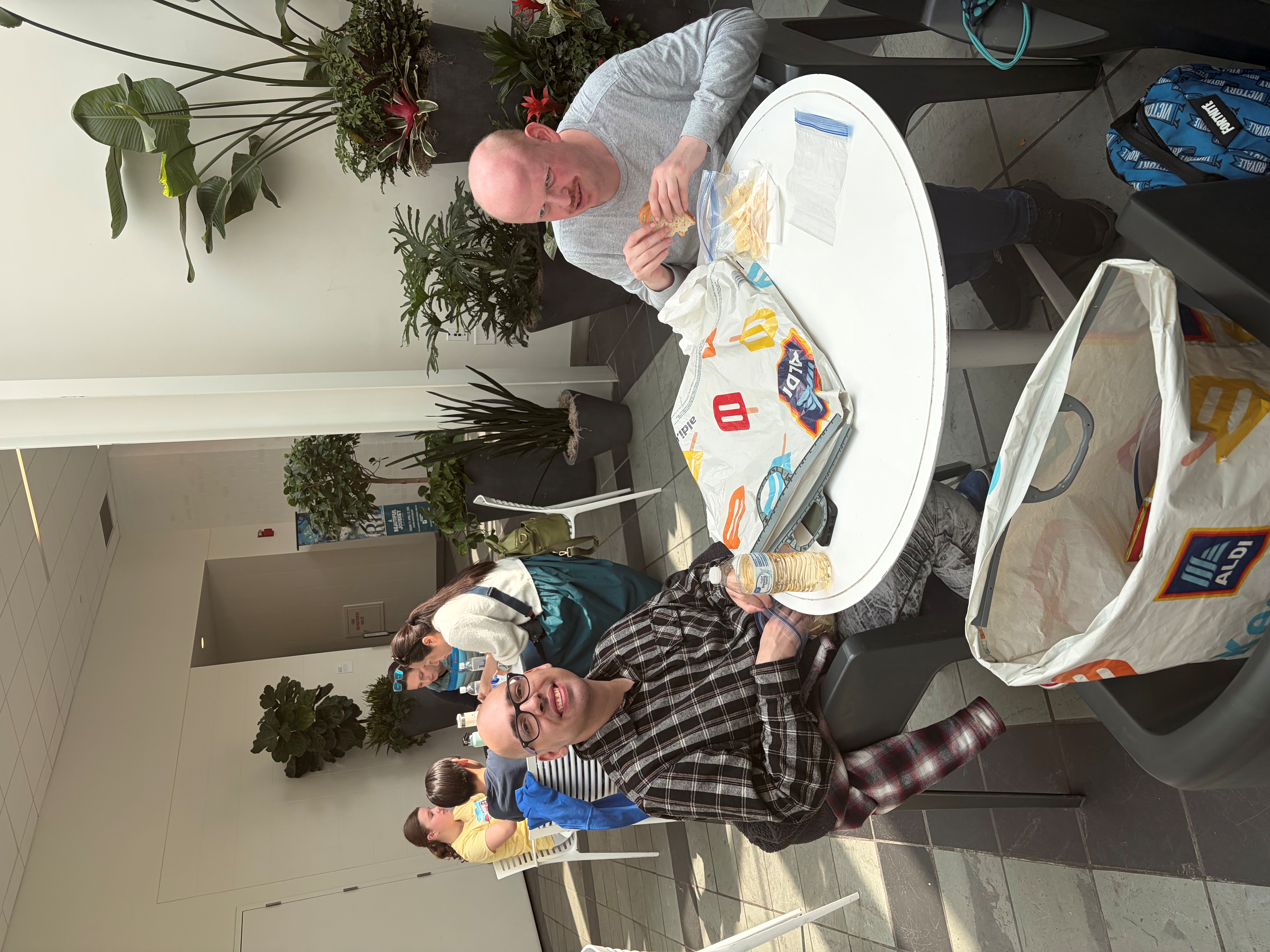 Two men sitting at a round white table eating sandwiches and chips in a bright indoor space with plants in the background.