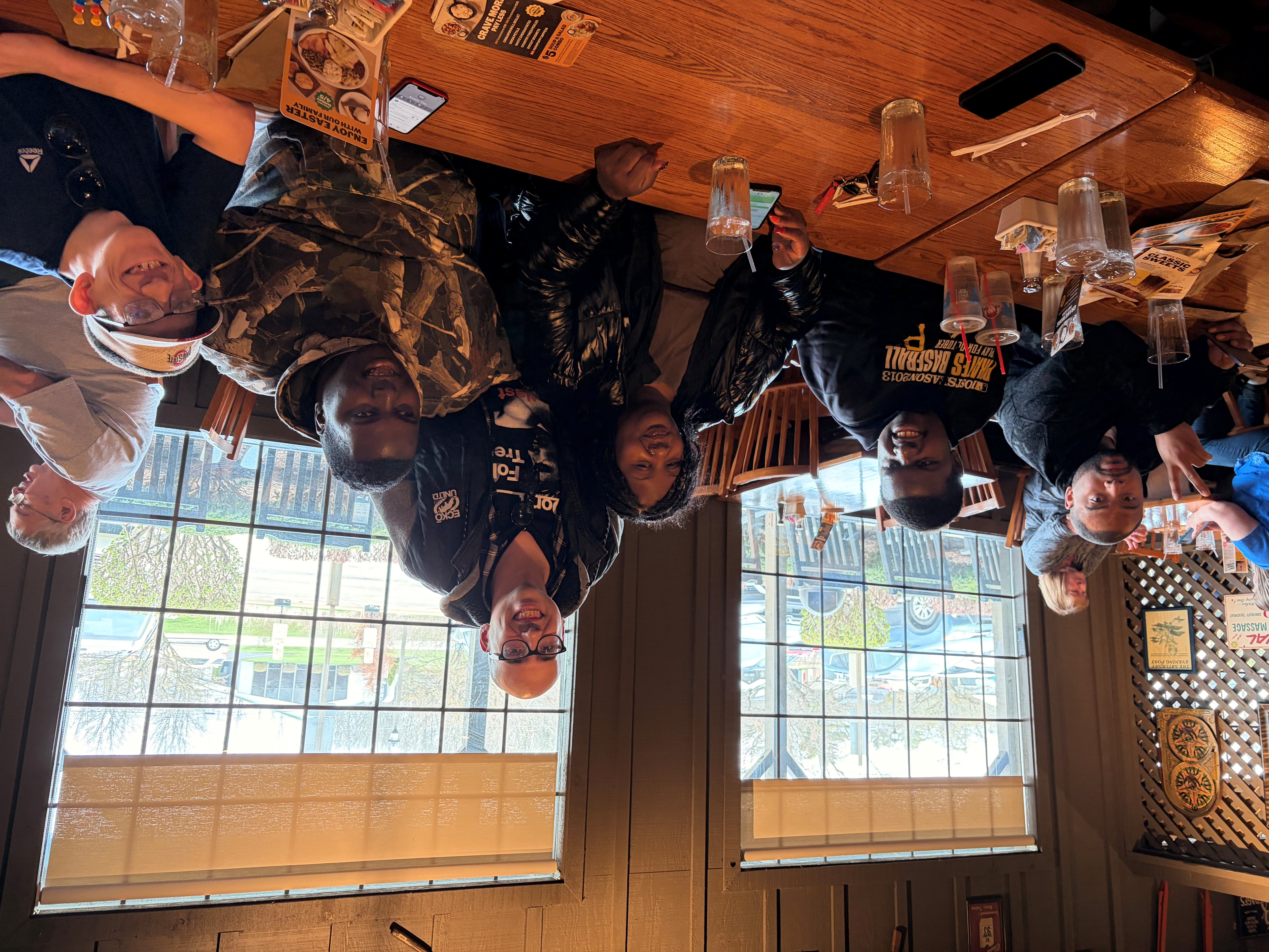 A group of six people smiling and sitting together at a wooden table in a restaurant with large windows showing an outdoor parking lot.