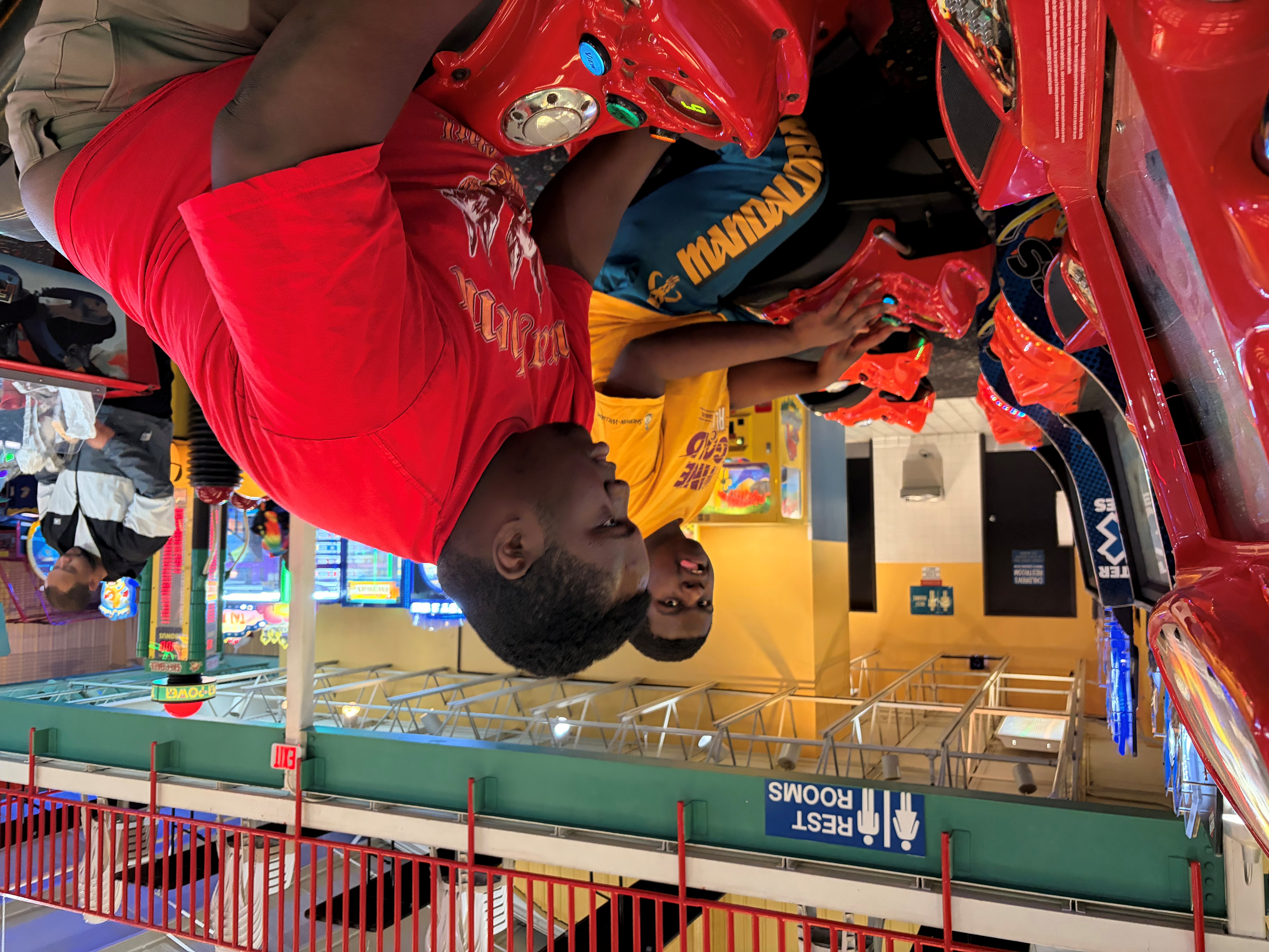 Two young men playing red arcade motorcycle racing games in an arcade center with a restroom sign visible above.