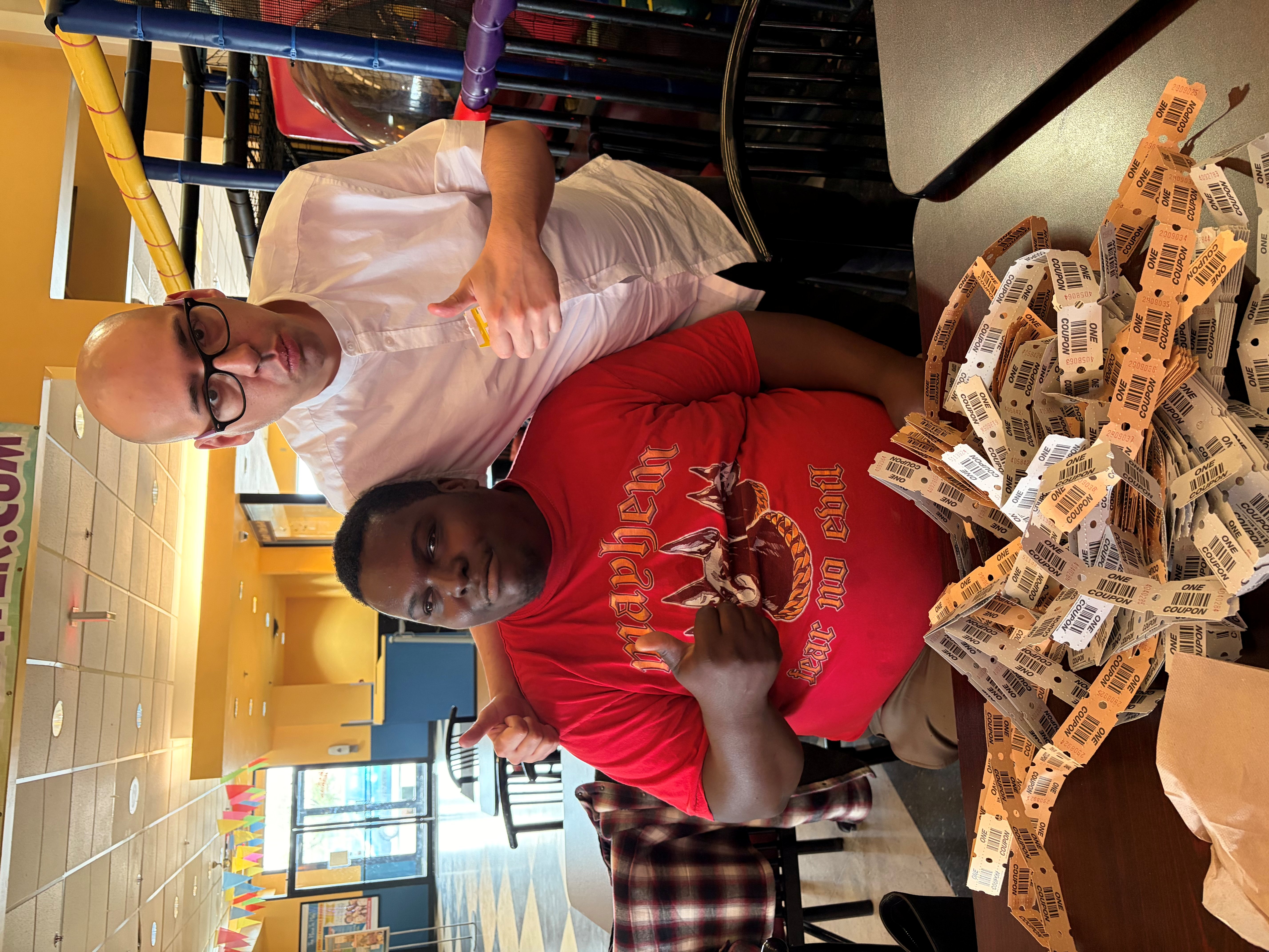 Two men sitting inside a restaurant giving thumbs up with a large pile of arcade or raffle tickets on the table in front of them.