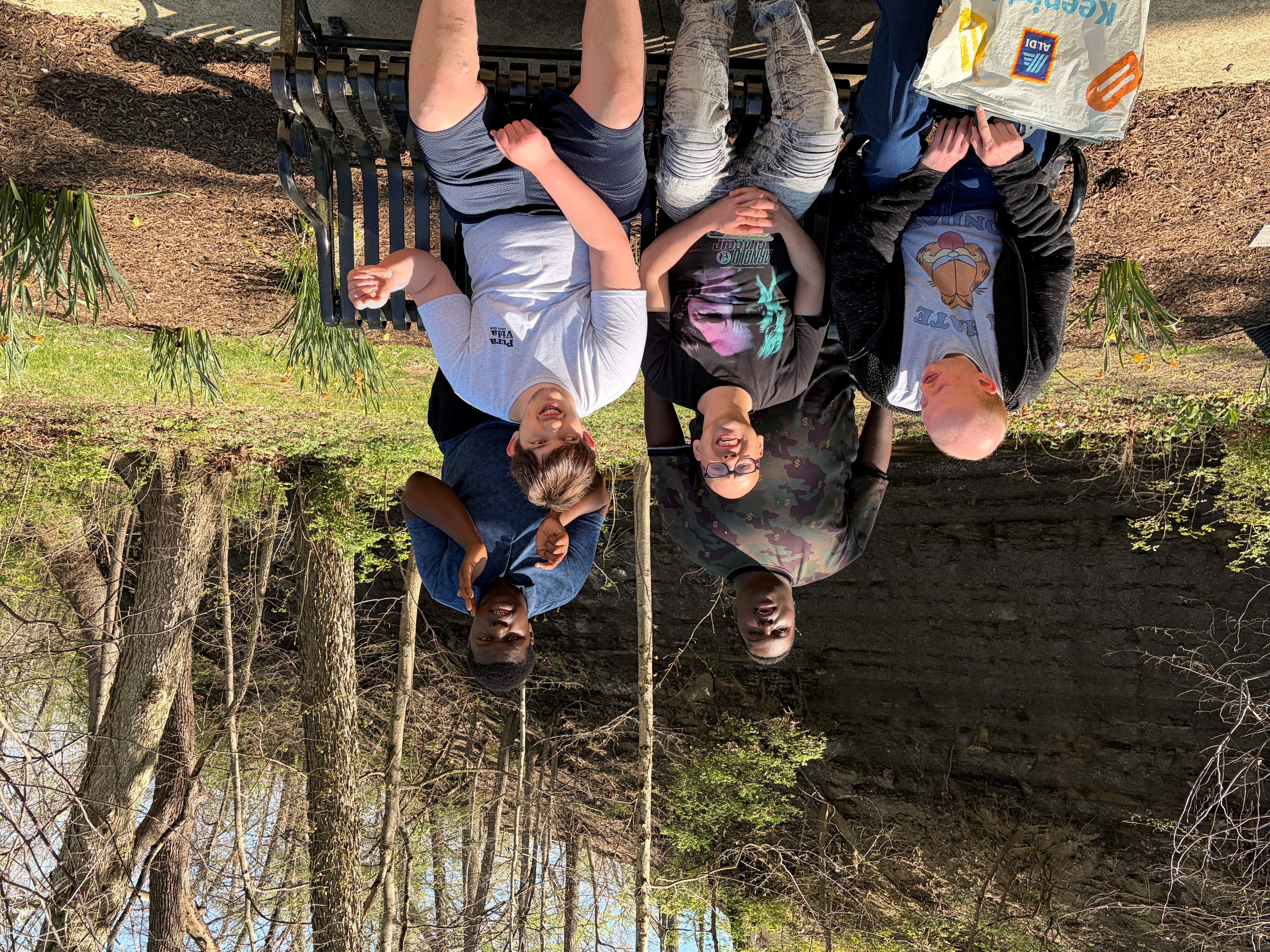 Group of five men outdoors, three sitting on a bench and two standing behind, surrounded by trees and greenery on a sunny day.
