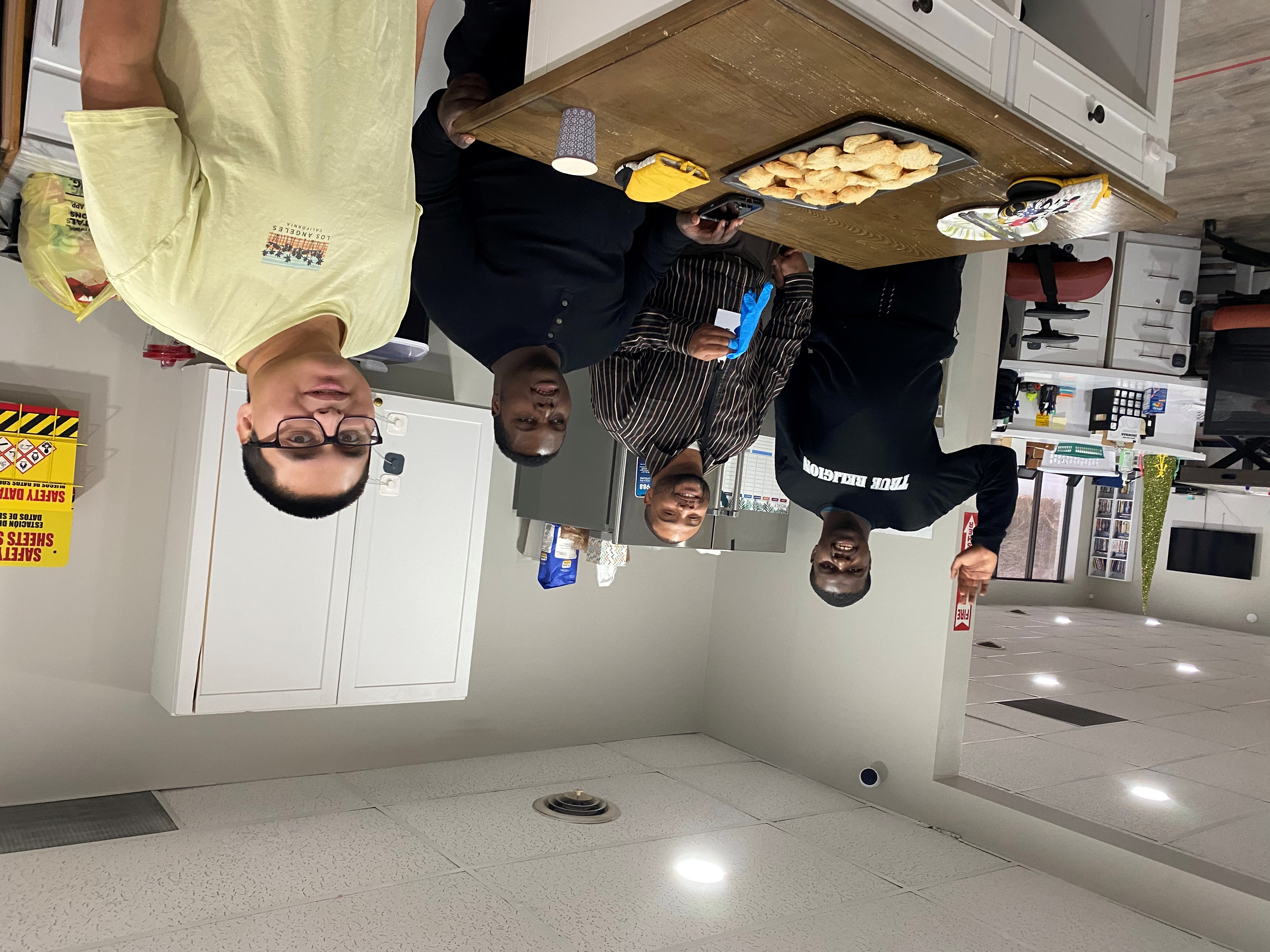 Four men standing in a kitchen area with a tray of baked goods on the counter in front of them.