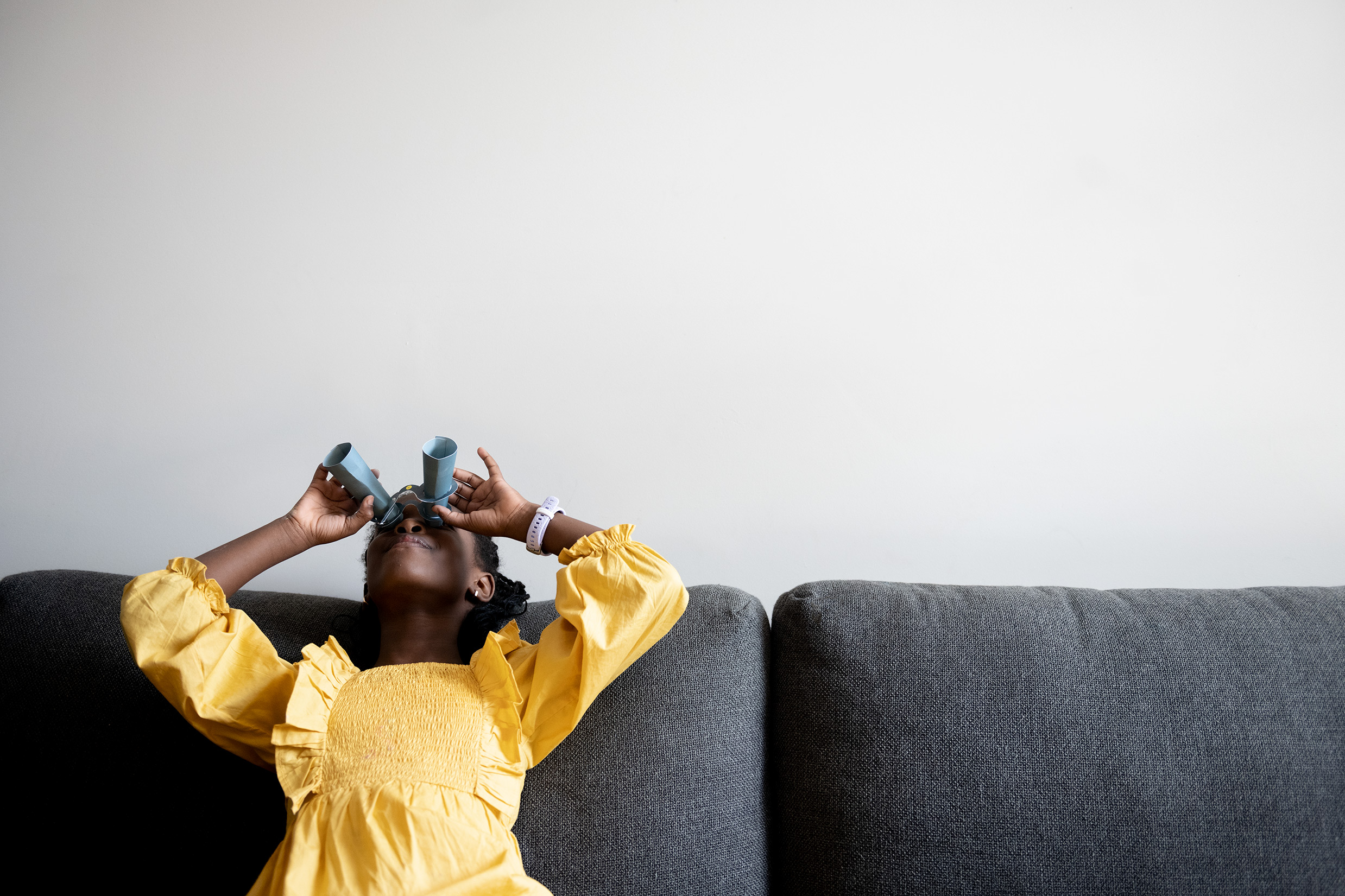 A young Black girl with braided hair leans back against a grey sofa, looking up through handmade blue cardboard binoculars. She is wearing a bright yellow ruffled dress against a plain, light-colored wall, capturing a moment of playful imagination.
