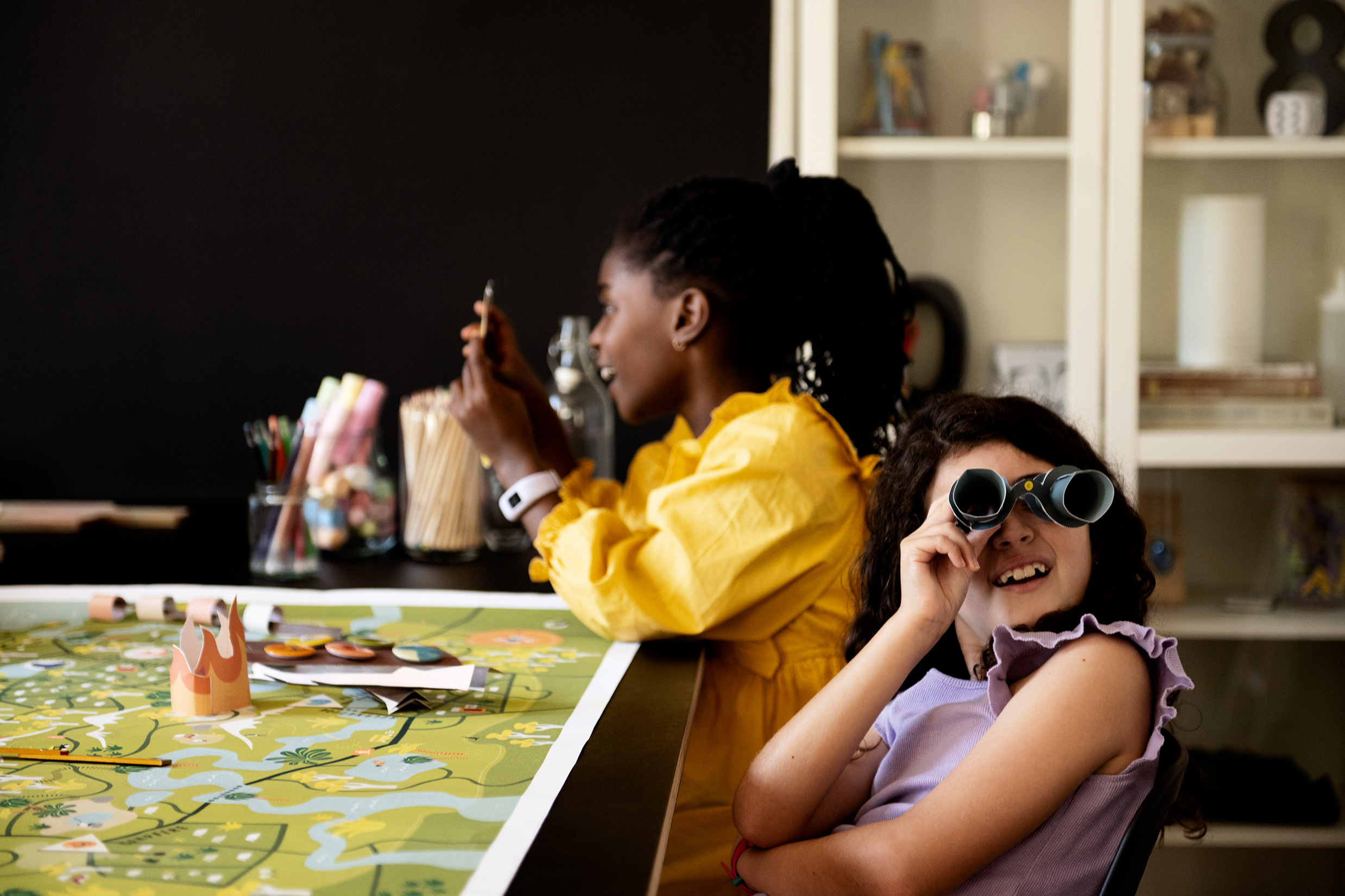 Two girls are engaged in a creative activity at a table. In the foreground, a girl in a purple ruffled top smiles while looking through blue toy binoculars. Beside her, the girl in the yellow dress works with craft materials. The table is covered with a la