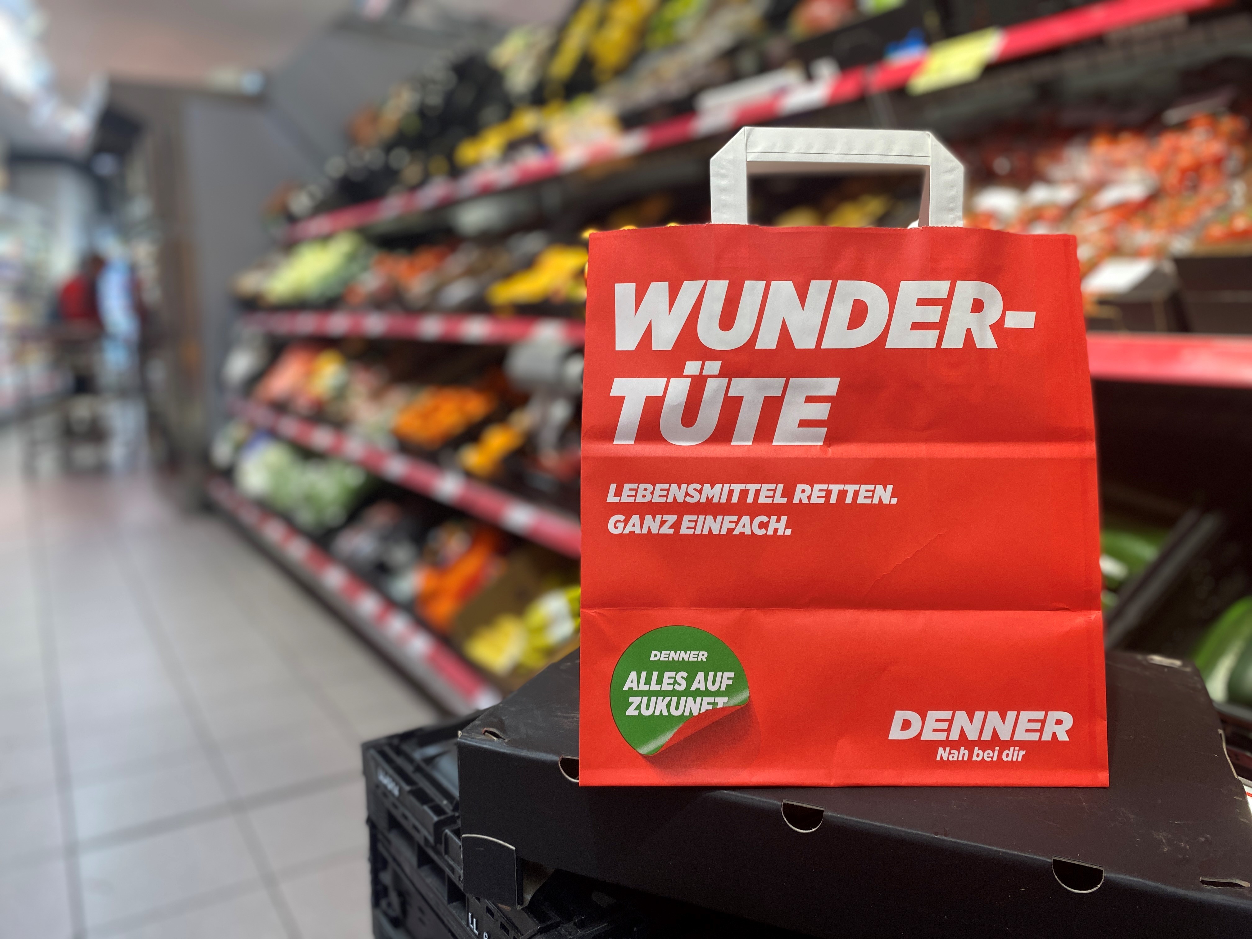 Eine Denner Wundertüte im Vodergrund, im Hintergrund eine Denner Filiale von innen und ein Regal mit Gemüse und Früchte darin