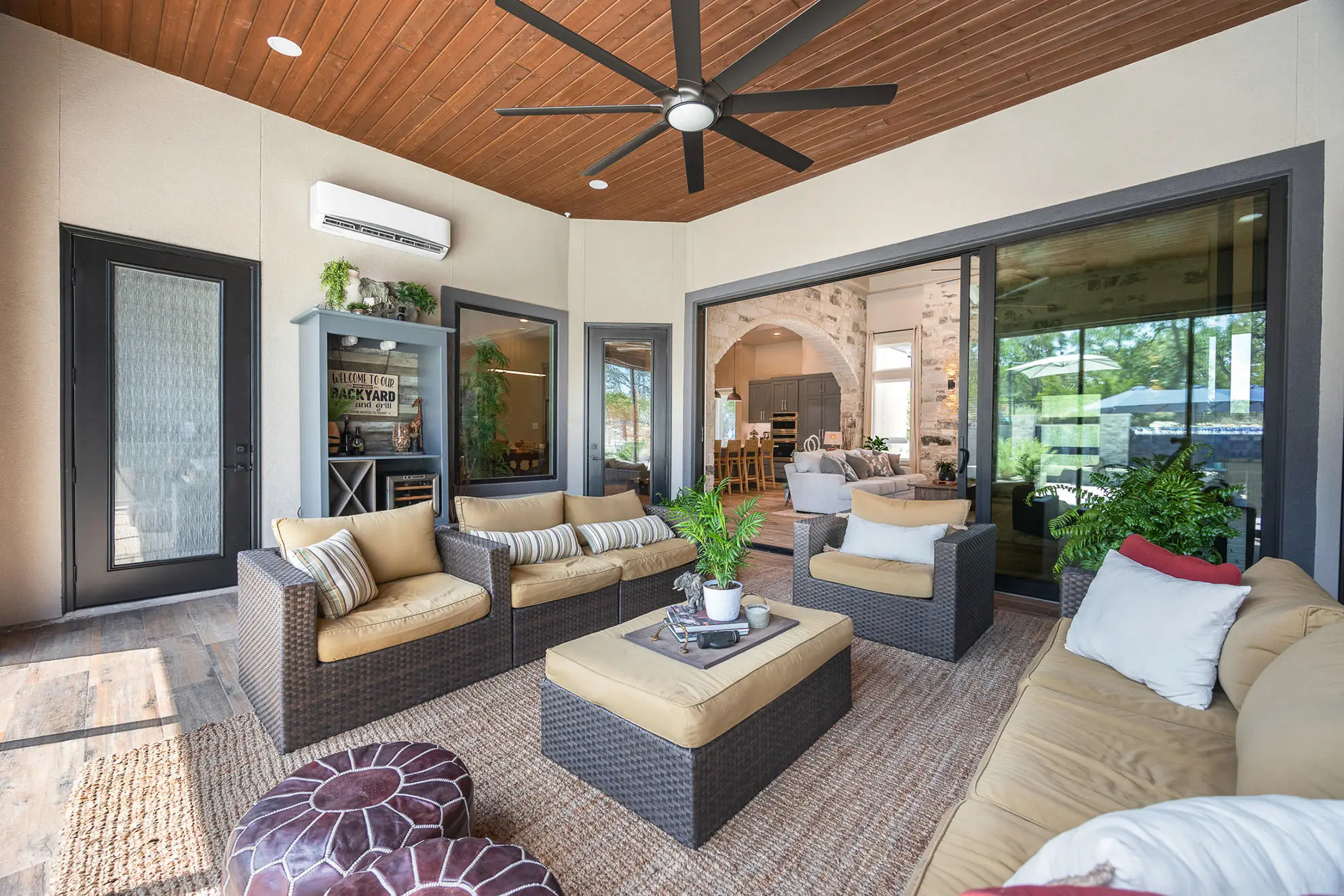 This stylish covered patio at a Cordillera Ranch home offers a relaxing outdoor retreat with cushioned wicker seating, wood ceiling, large fan, and seamless indoor-outdoor flow into a spacious living room with stone accents and high-end finishes—crafted by KC Custom Homes.