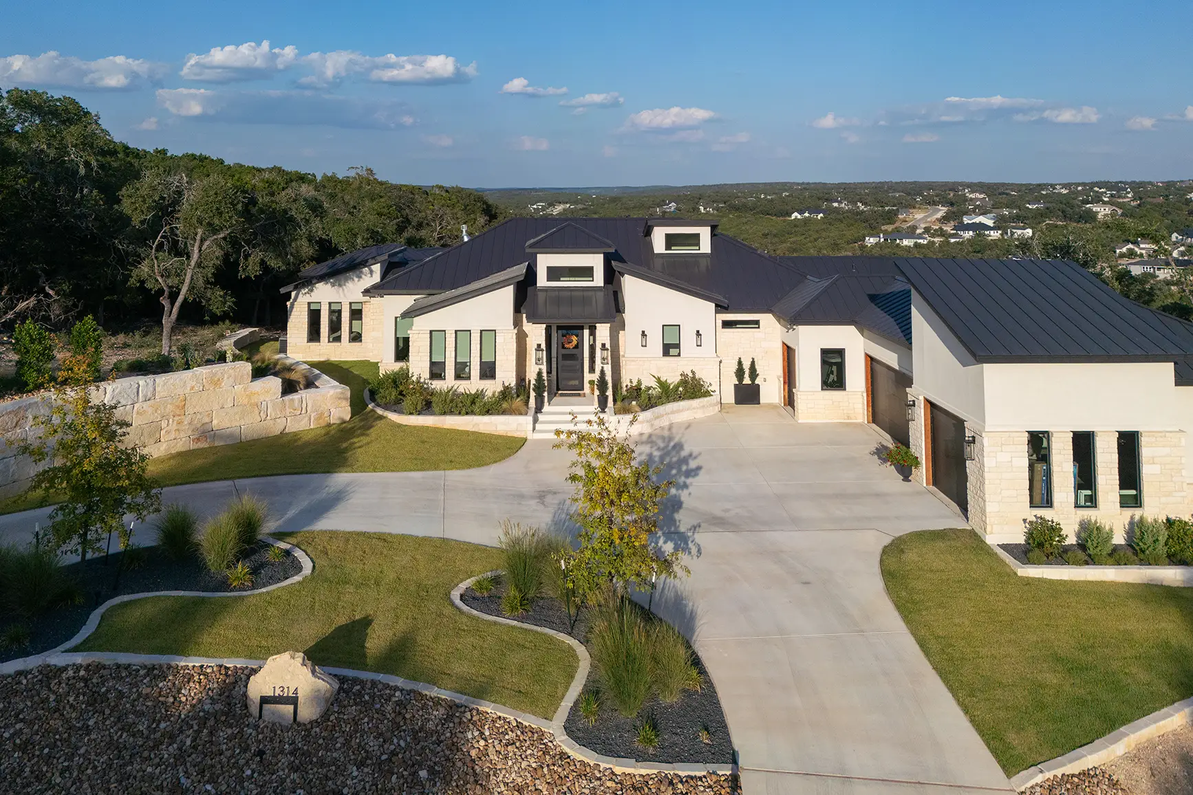 A stunning aerial view of a modern farmhouse-style custom home in Cordillera Ranch, featuring a black metal roof, light stone facade, beautifully landscaped circular driveway, and panoramic views of the Texas Hill Country—designed by KC Custom Homes.