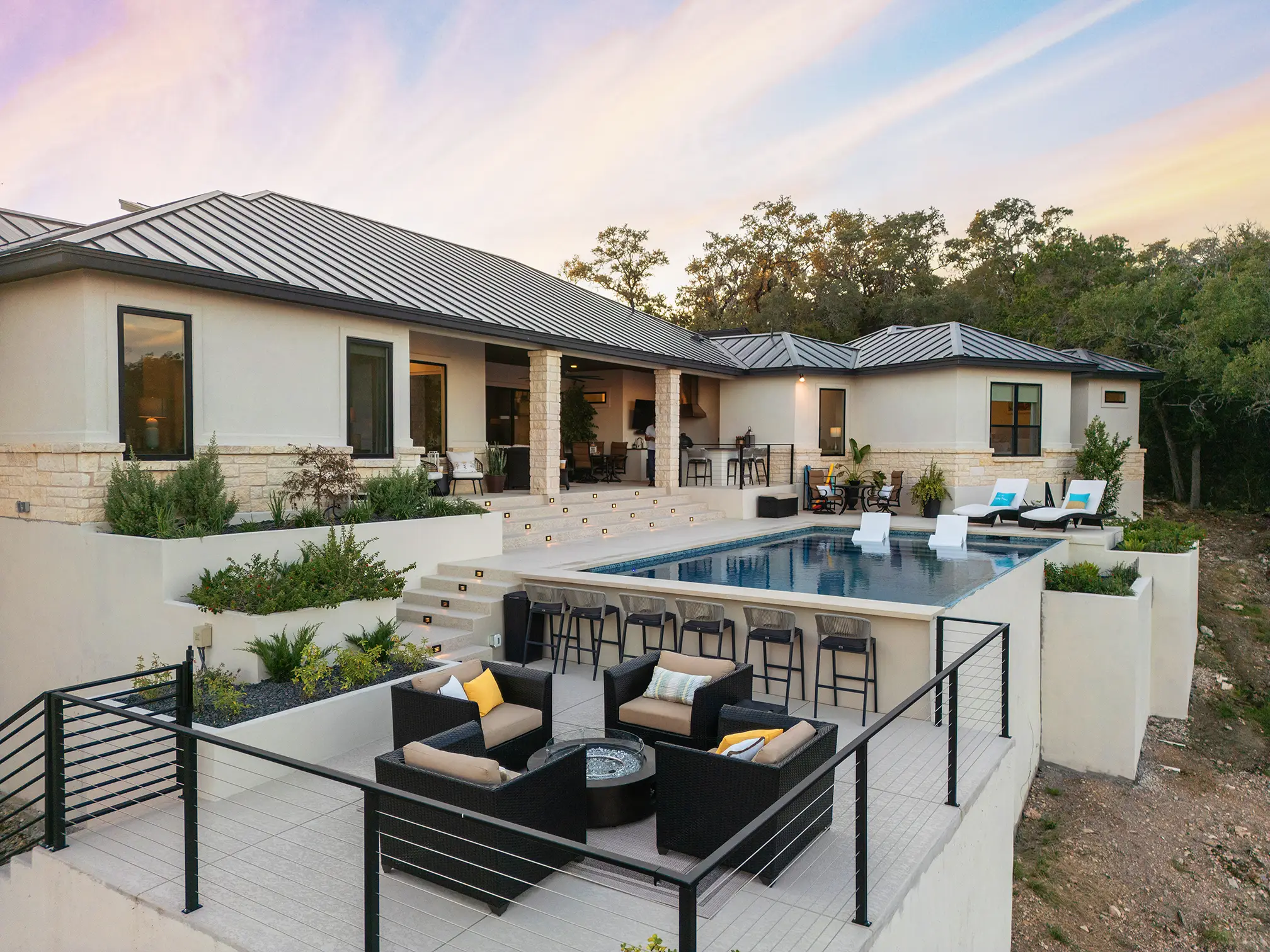 A breathtaking view of a luxury custom home in Cordillera Ranch at sunset, showcasing an infinity-edge pool, contemporary outdoor furniture, and a multi-tiered patio designed for upscale entertaining—crafted by KC Custom Homes in Boerne, TX.