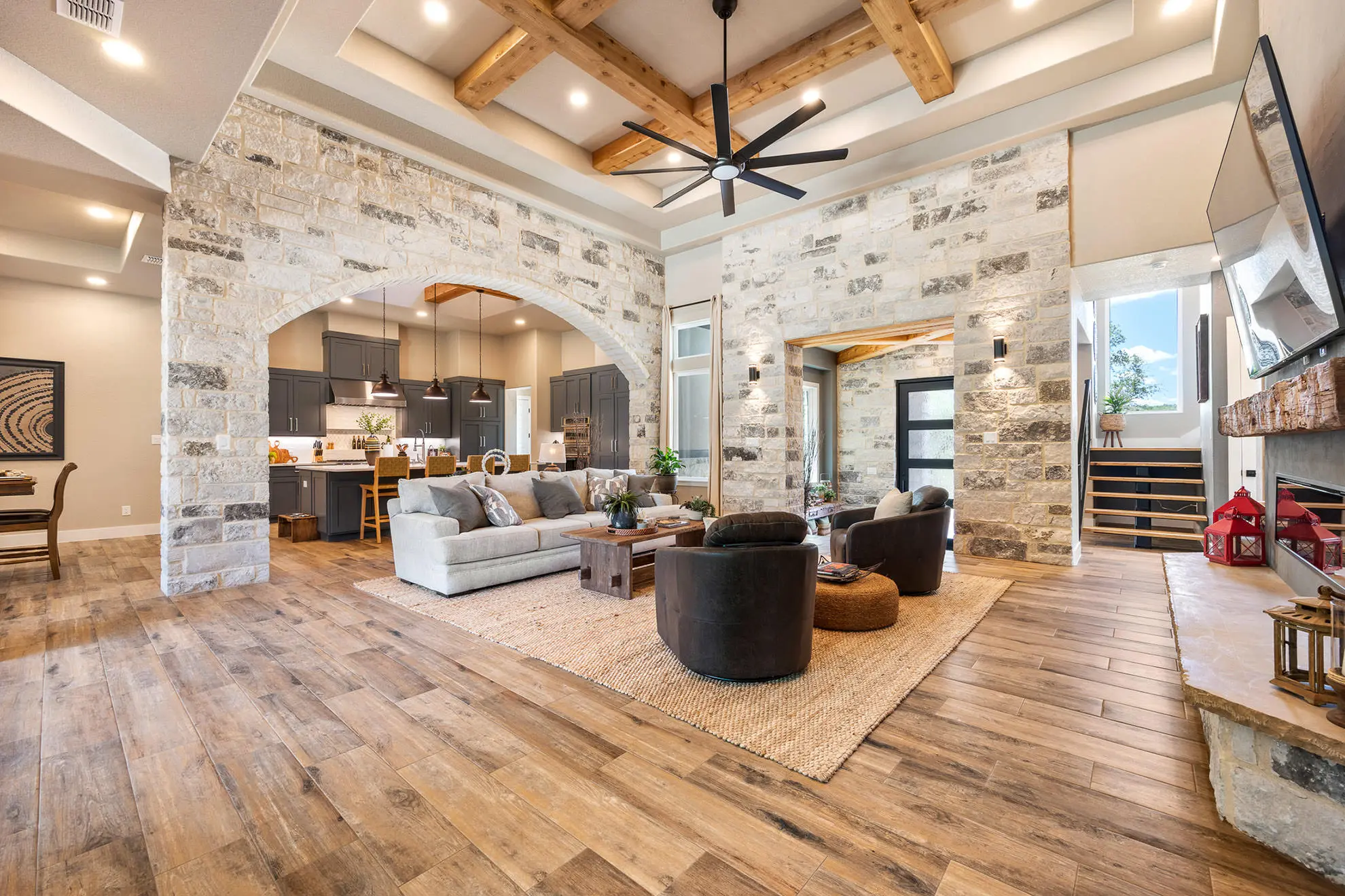 This luxurious open-concept living room in Cordillera Ranch showcases a blend of rustic charm and modern elegance with arched stone walls, exposed wood beams, a floor-to-ceiling fireplace, and seamless connection to the kitchen—highlighting KC Custom Homes’ signature Hill Country craftsmanship.