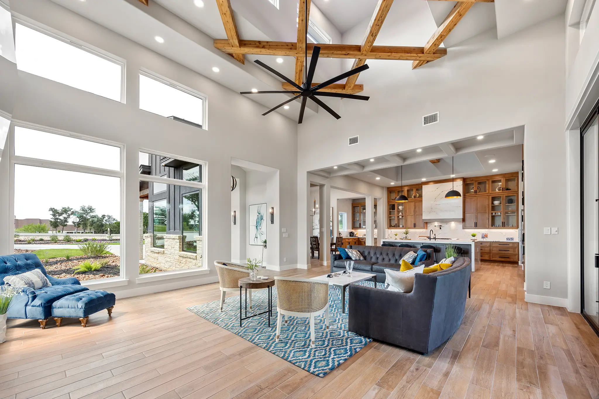 Open-concept Hill Country living room with vaulted wood-beam ceiling, large windows, and seamless flow to the kitchen by KC Custom Homes