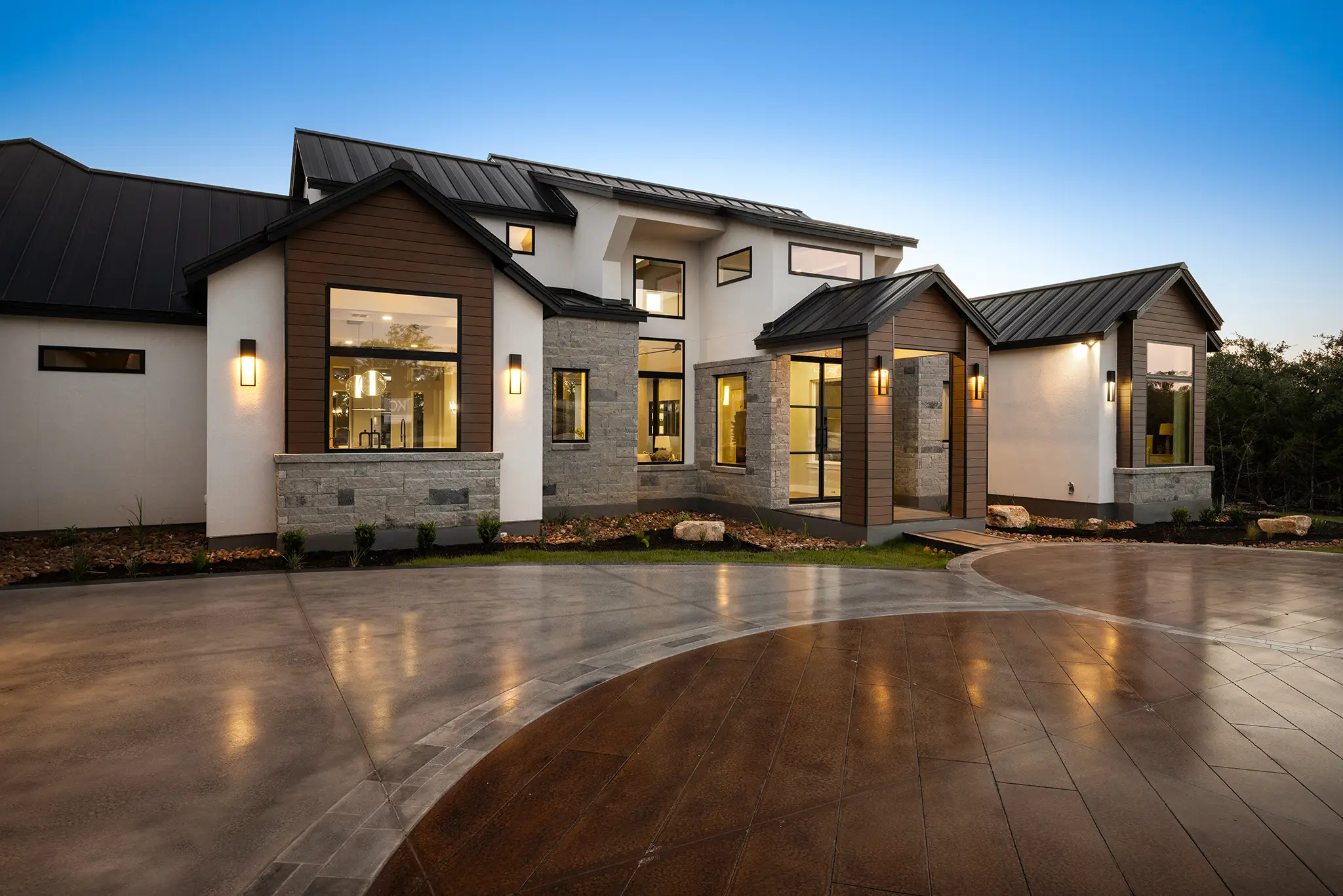Modern Hill Country custom home by KC Custom Homes at dusk, featuring a metal roof, large windows, stone and wood accents, and a curved driveway in New Braunfels Texas.