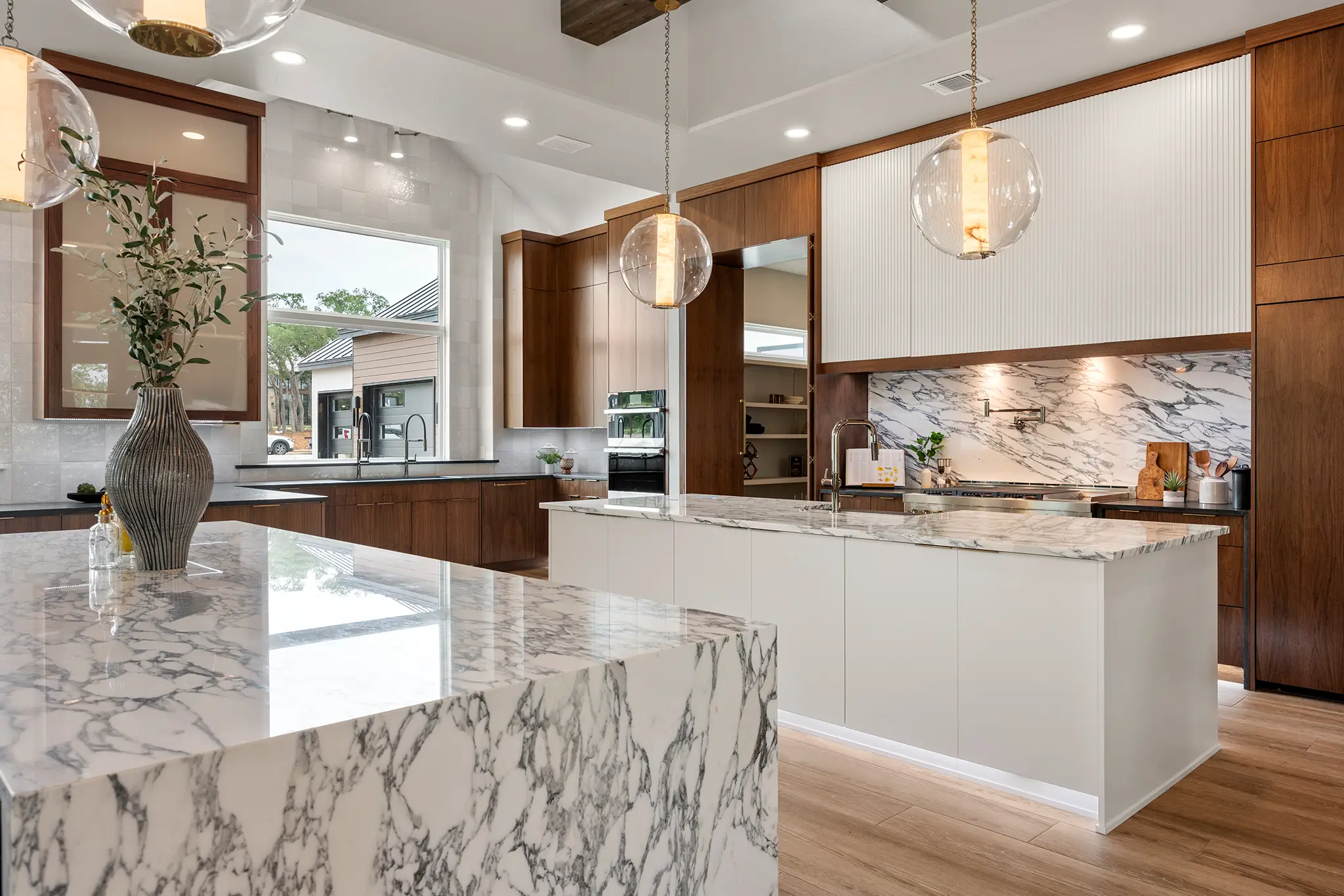 Luxury kitchen in a KC Custom Homes residence with a marble waterfall island, warm wood cabinetry, pendant lighting, and a large window overlooking the property in New Braunfels Texas.