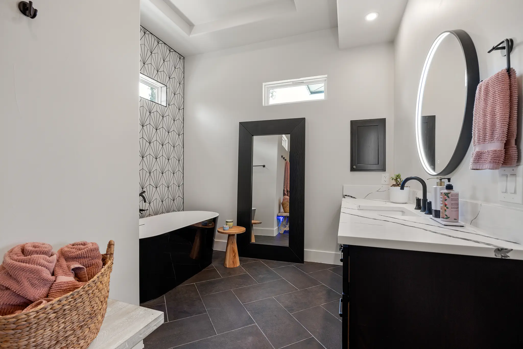 Spacious modern bathroom with a freestanding tub, patterned tile accent wall, large framed mirror, and dual-sink vanity in a custom home in Mitchell Ranch, Bulverde Texas.